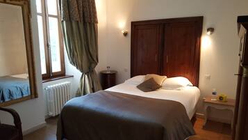 A bedroom with a bed, a radiator, a mirror, a window with curtains, a bedside table with a bottle, and a clock.