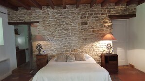 Un dormitorio con pared de piedra, una cama con ropa de cama blanca, dos mesitas de noche con lámparas y techo de madera.