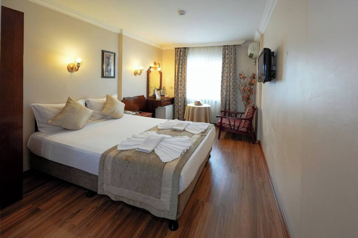 A hotel room with a large bed, bedside tables, a small table with a chair, and a TV mounted on the wall.