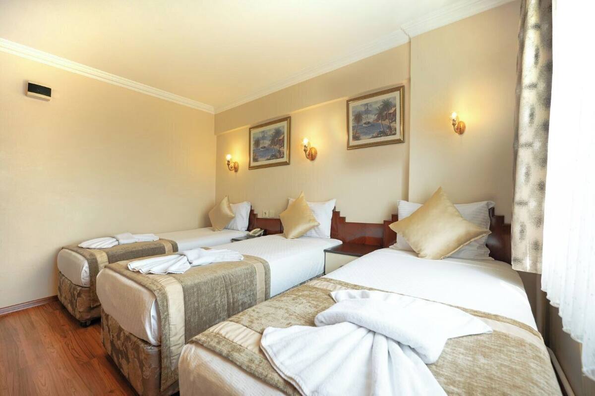 A hotel room with four beds arranged in a row, each with white linens and beige headboards. There are wall-mounted light fixtures and framed pictures above the beds.