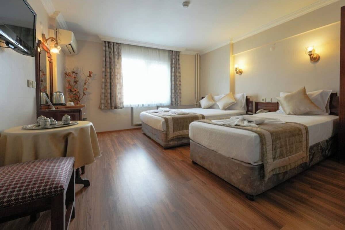 A hotel room with two beds, a dining table, a bench, and a large window with curtains.
