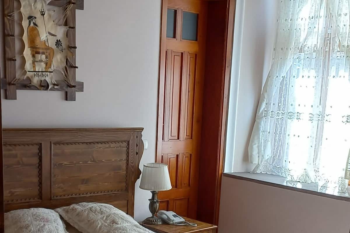 Ein Schlafzimmer mit hölzernem Kopfteil, einem Bett mit weißer Bettdecke, einem Nachttisch mit Lampe, einer Holztür und einem Fenster mit durchsichtigem Vorhang.
