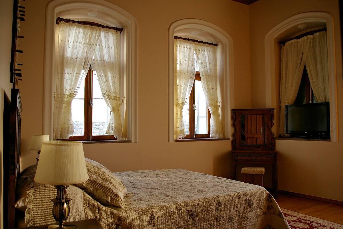 Ein Schlafzimmer mit einem Bett, einer Lampe, einem Fernseher und zwei mit Vorhängen versehenen Fenstern.