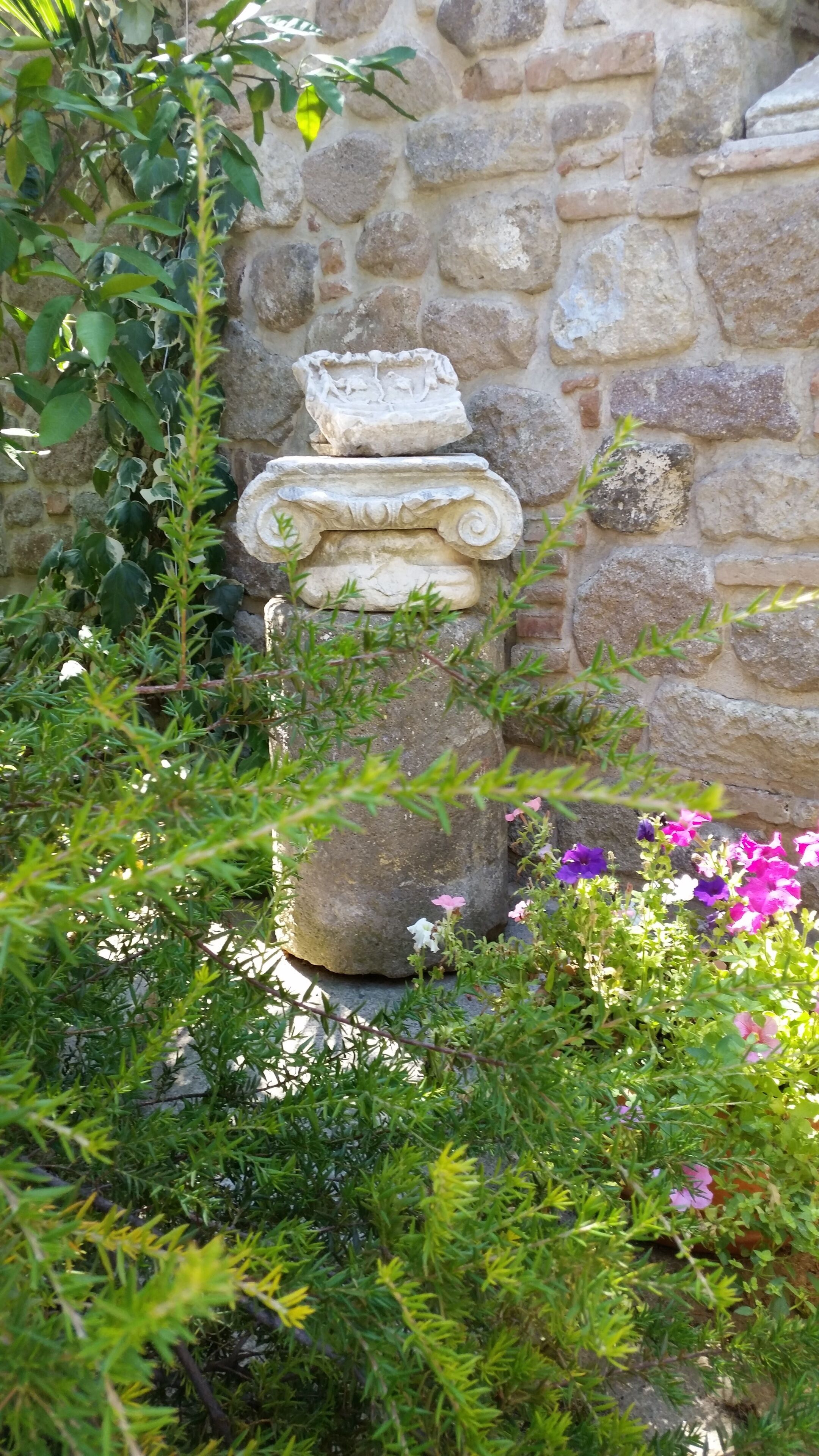 Un muro de piedra con una estructura decorativa de piedra, vegetación exuberante y flores moradas.