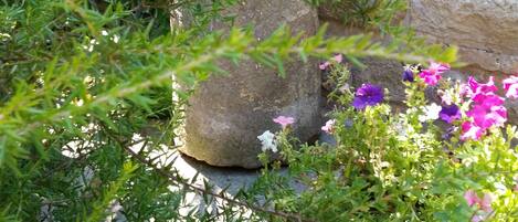 Un muro de piedra con una estructura decorativa de piedra, vegetación exuberante y flores moradas.