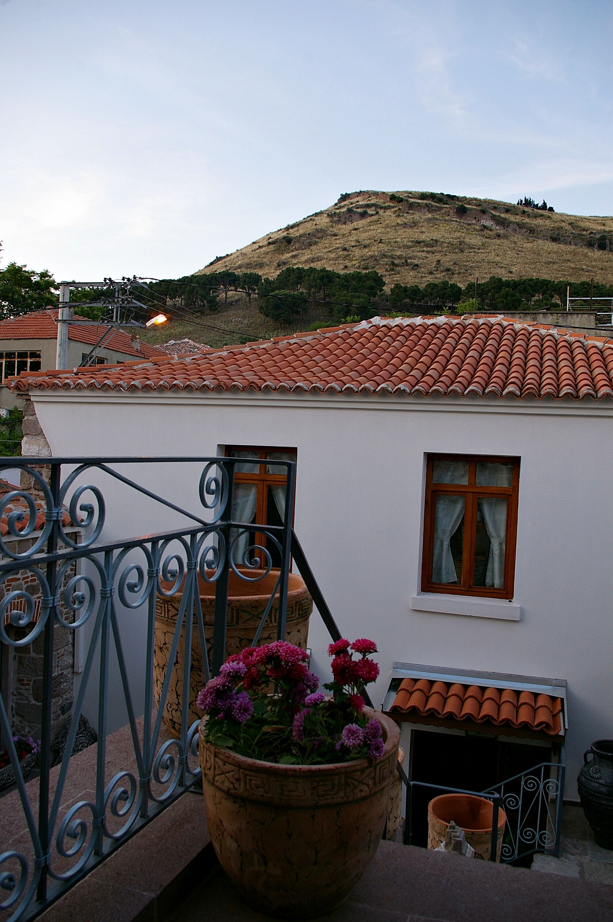 A white building with a red-tiled roof, a balcony with a wrought iron railing, and a large terracotta pot with flowers.