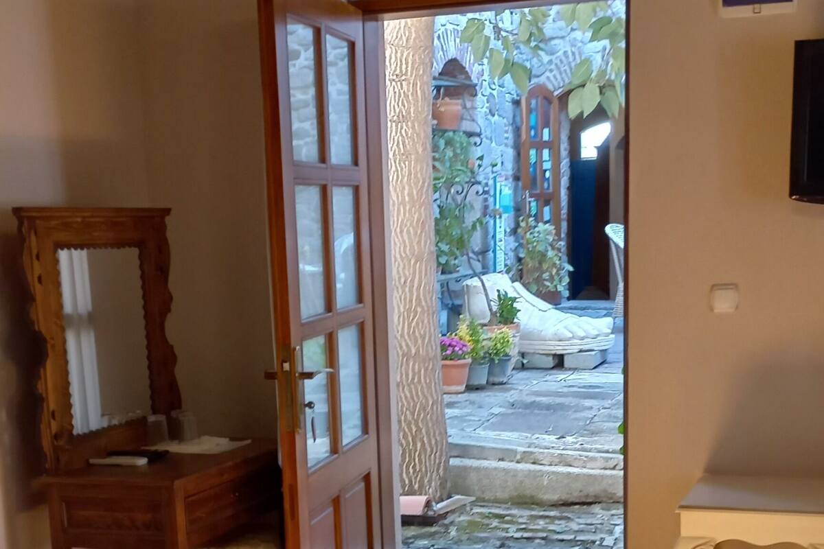 Ein Schlafzimmer mit einem hölzernen Bett, einer Holzkommode, einem Spiegel, einem weißen Nachttisch und einer geöffneten Tür, die zu einem Innenhof mit Pflanzen und einer Bank führt.
