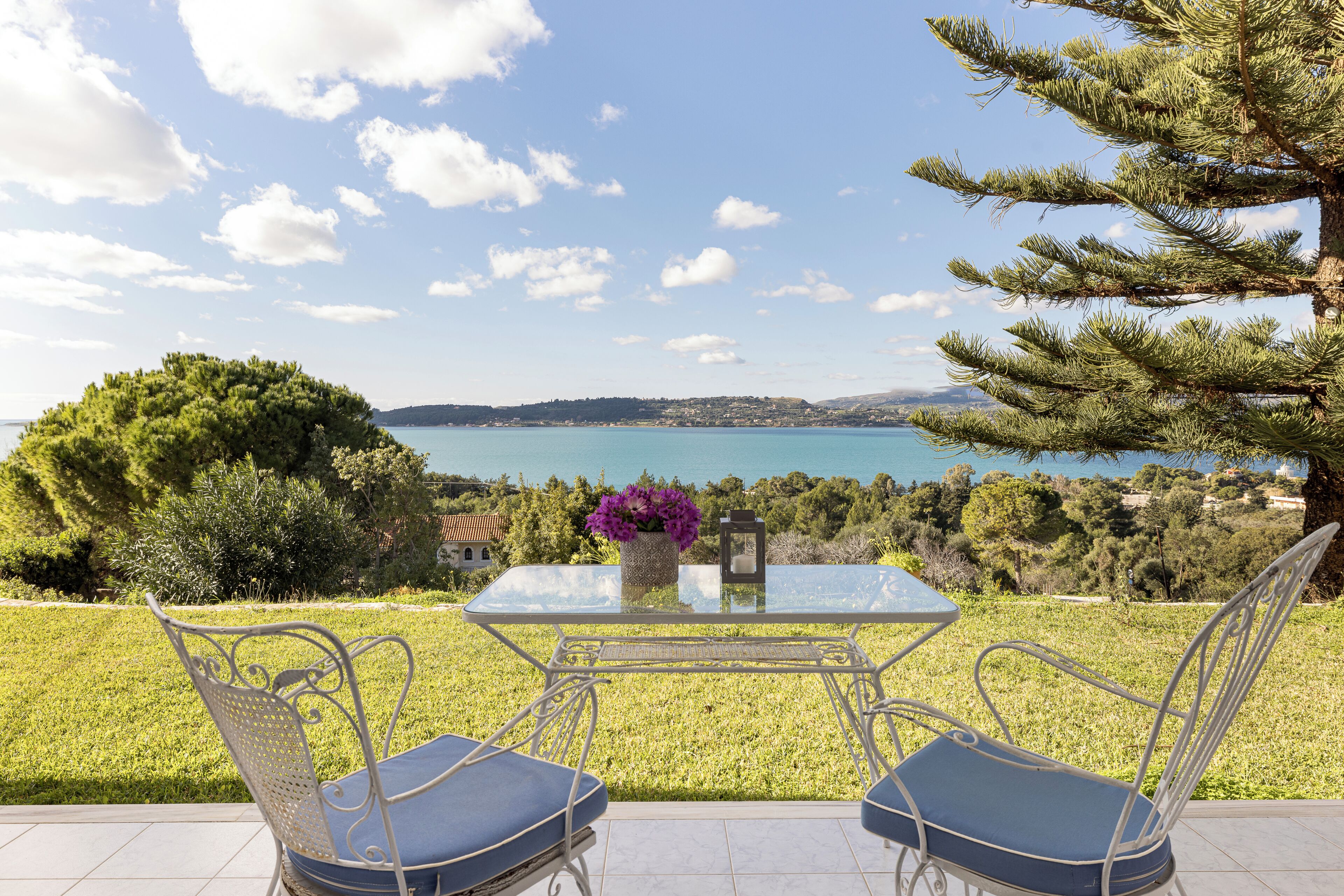 Une terrasse avec une table et deux chaises, offrant une vue panoramique sur un plan d’eau et des collines.