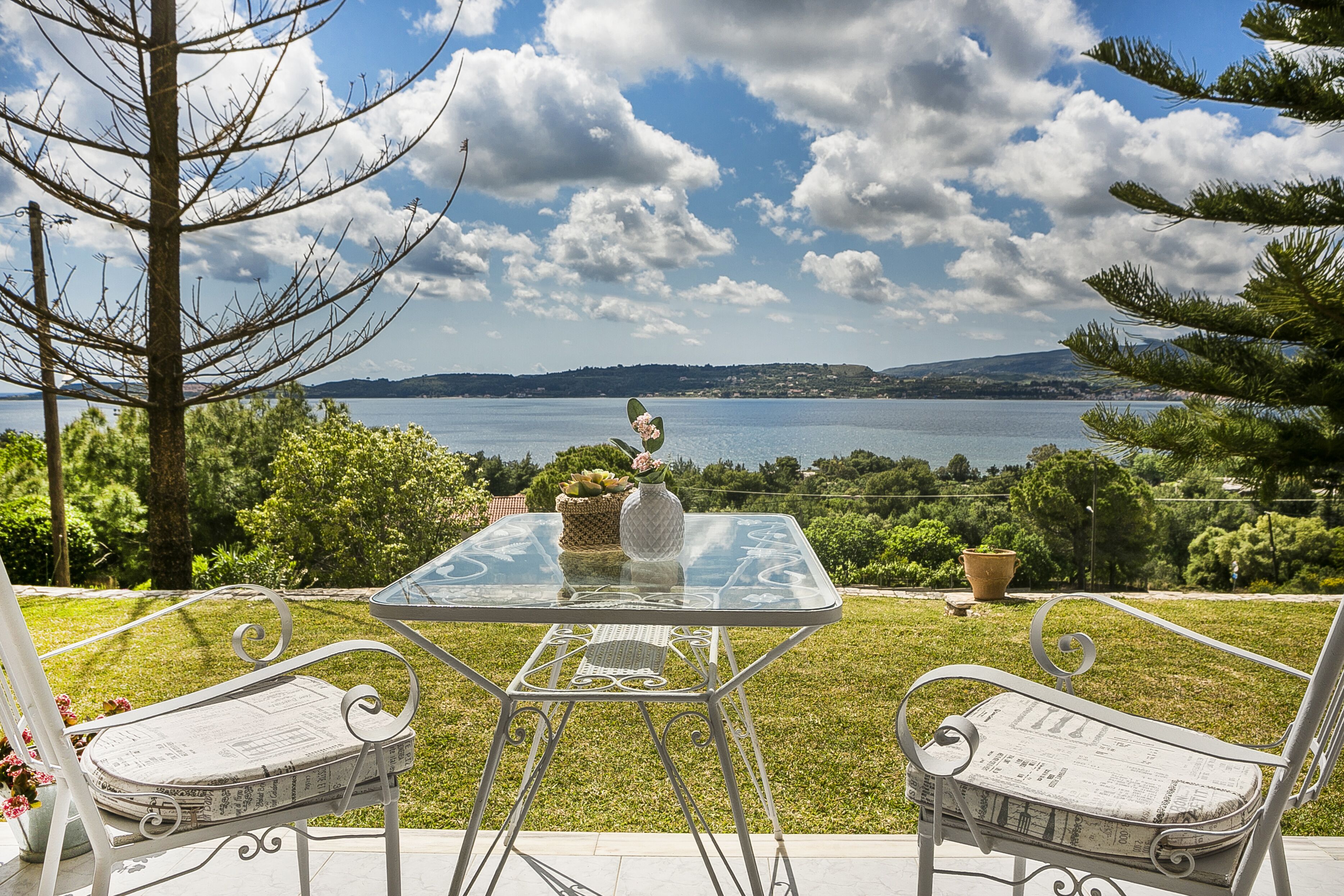 A glass table with two white chairs, a vase with flowers, and a view of a lake and hills.