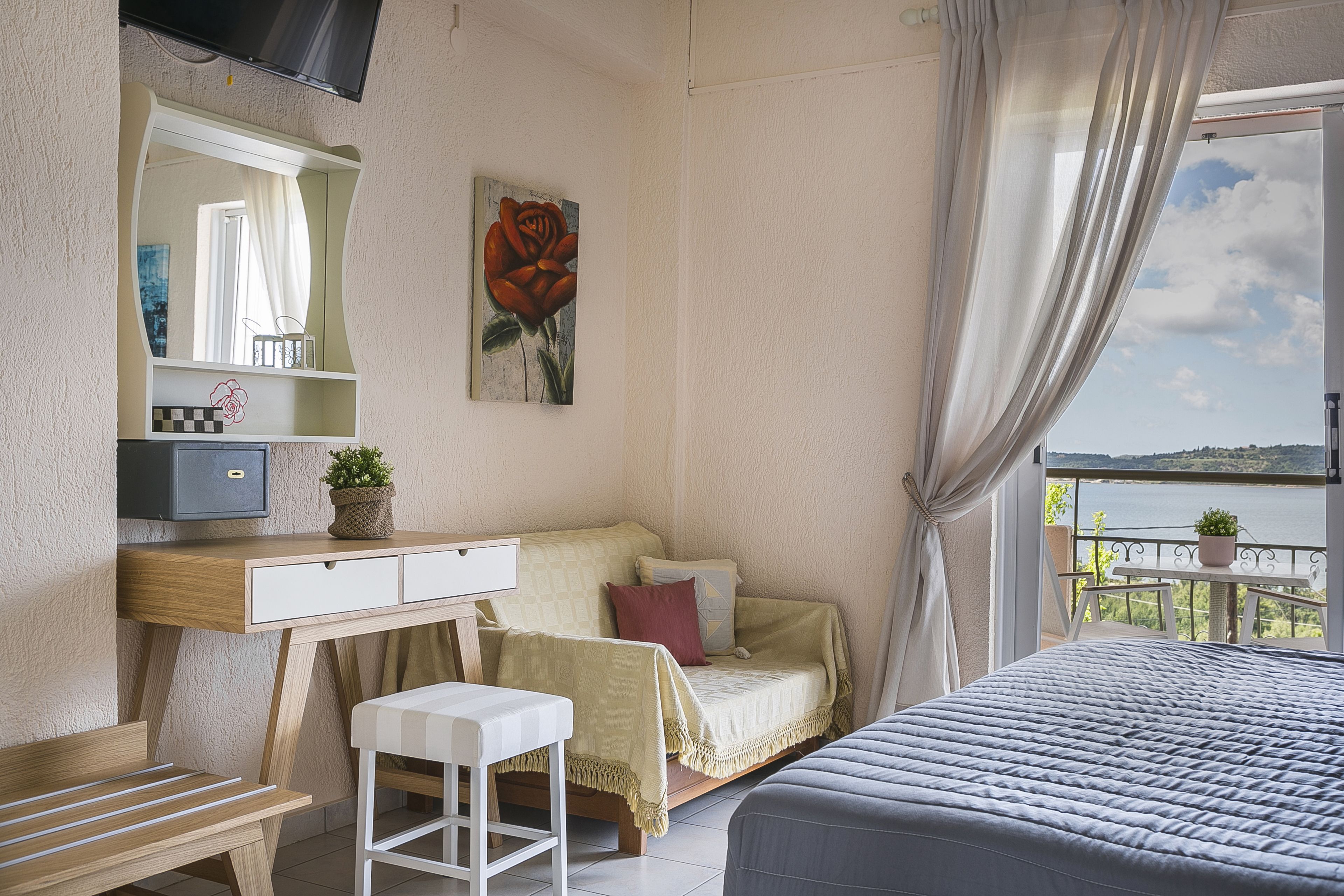 A bedroom with a bed, a desk, a chair, a bedside table, a mirror, a window with curtains, and a view of the outdoors.