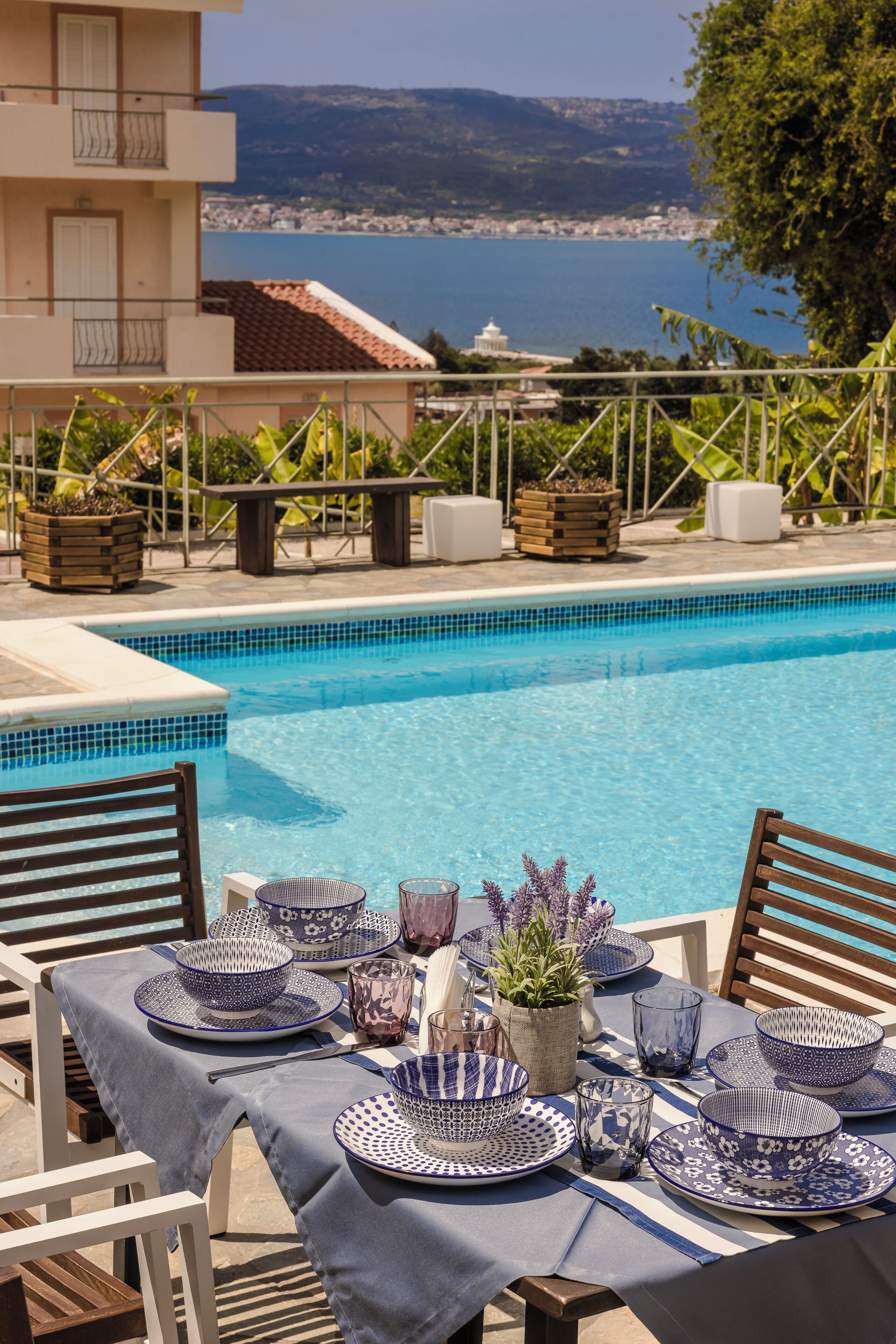 A poolside dining area with a table set for two, overlooking a body of water and a mountainous landscape.