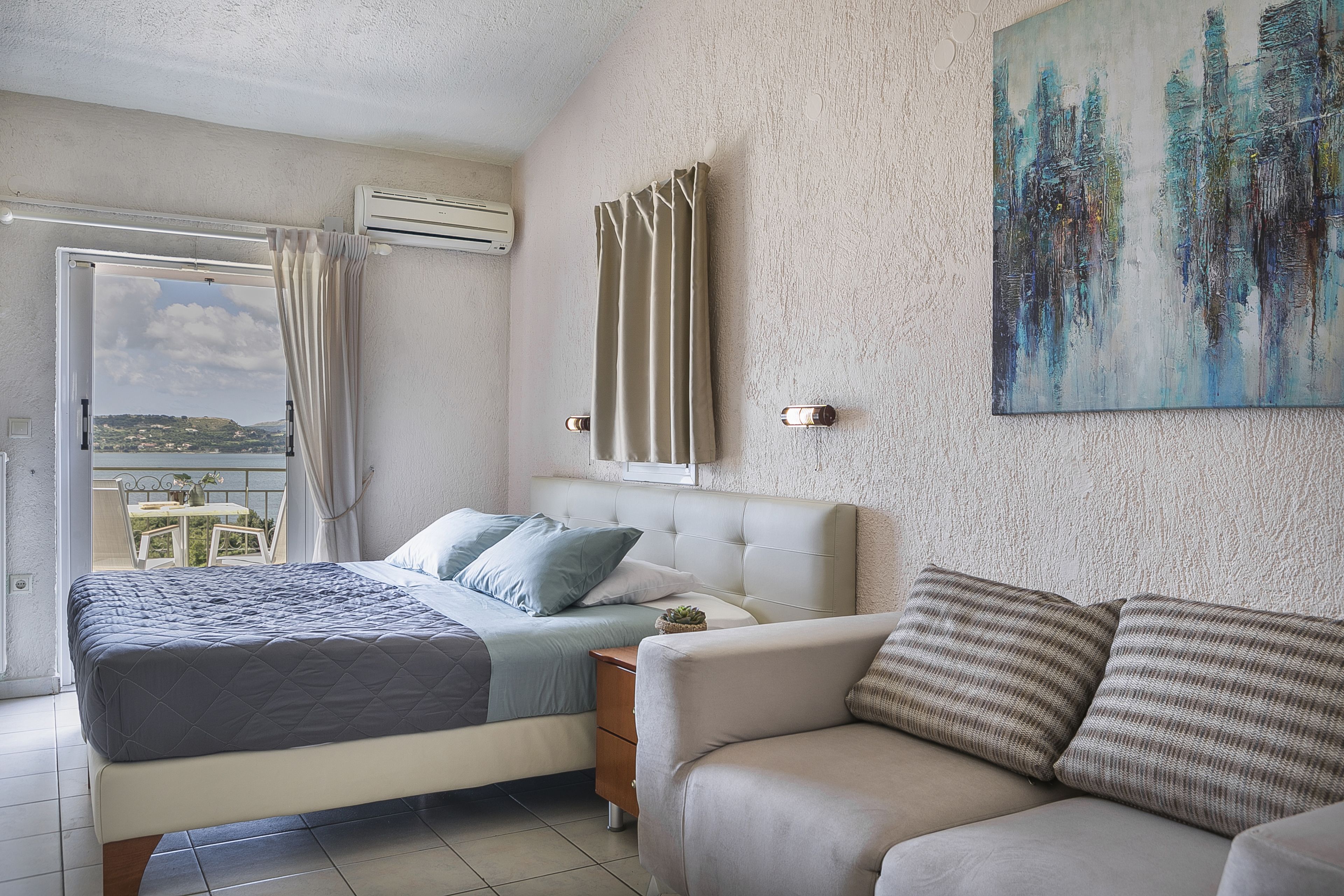 Un dormitorio con cama, sofá, aire acondicionado, ventana con cortinas y un cuadro en la pared.