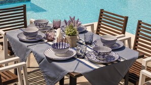 Un espace repas aménagé au bord de la piscine, avec une table dressée pour deux personnes, offrant une vue sur une étendue d’eau et une côte lointaine.