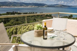 A round glass table with a bottle of wine, glasses, and a woven basket on a balcony overlooking a coastal landscape. - Panorama Fanari Studios & Apartments (Kefalonia)