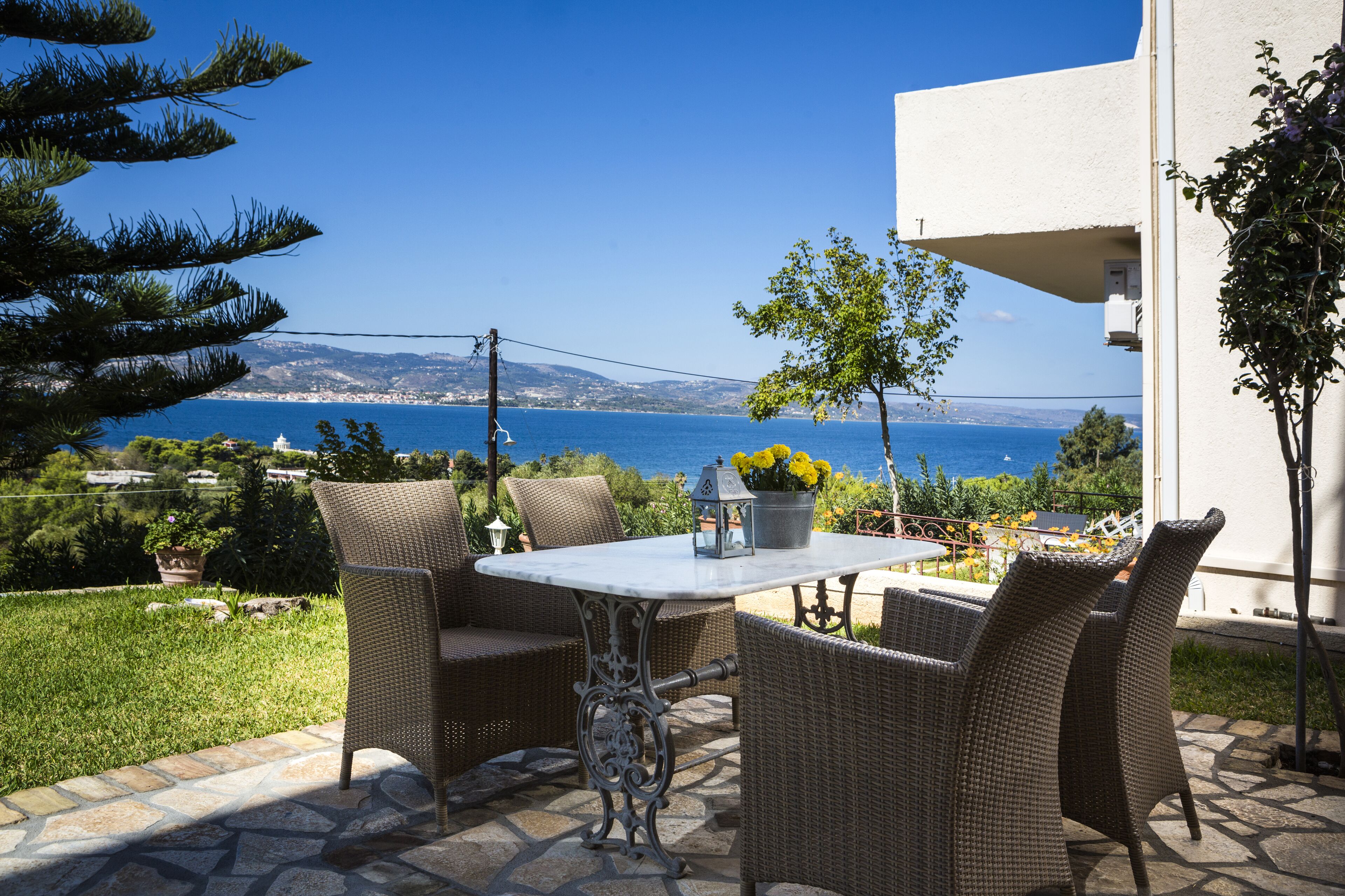 An outdoor patio with wicker furniture, a table with a vase of yellow flowers, and a view of the sea and hills in the background.