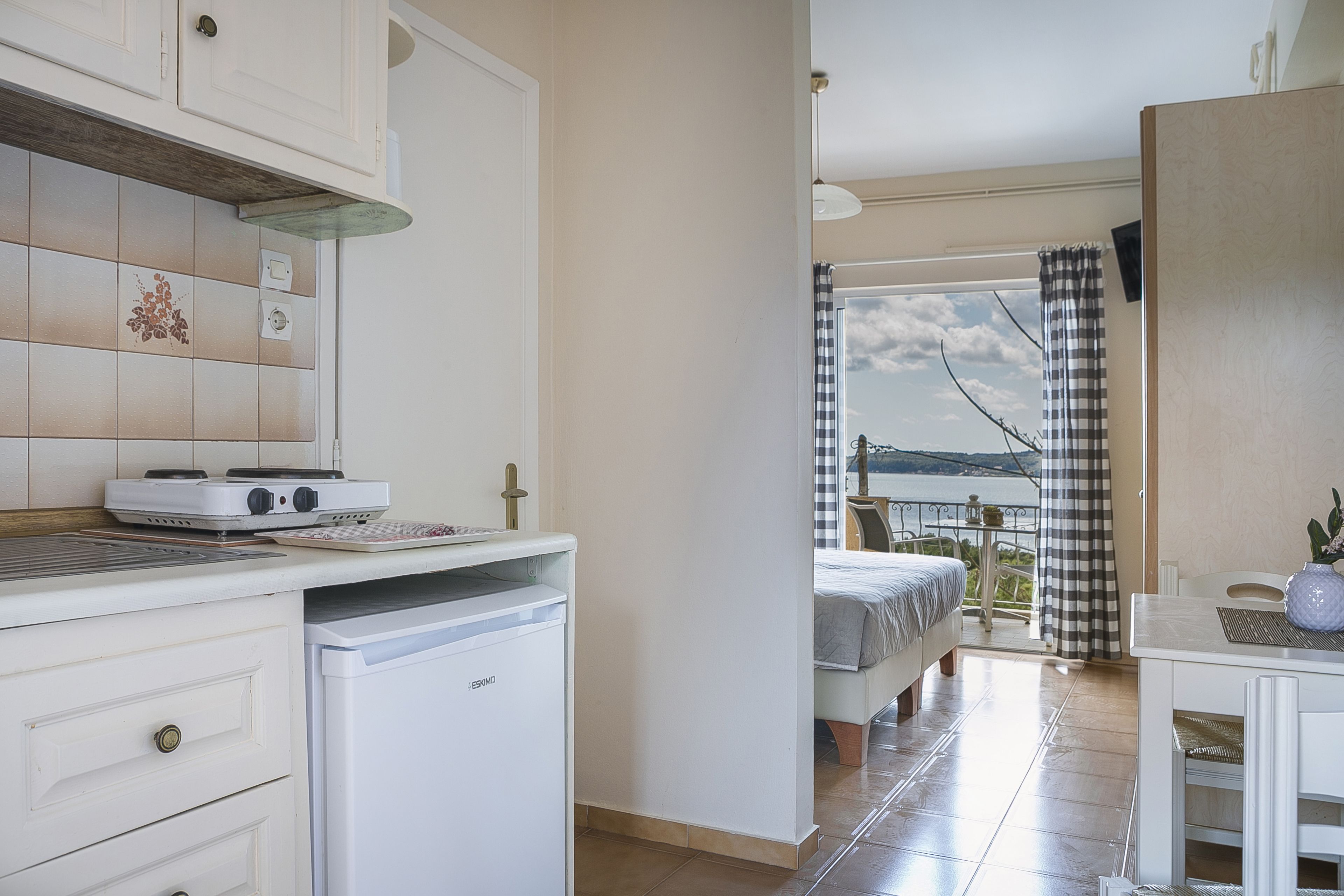 Cocina pequeña con estufa, refrigerador y zona de comedor. Se ve un balcón con cortina a cuadros y vista al mar.