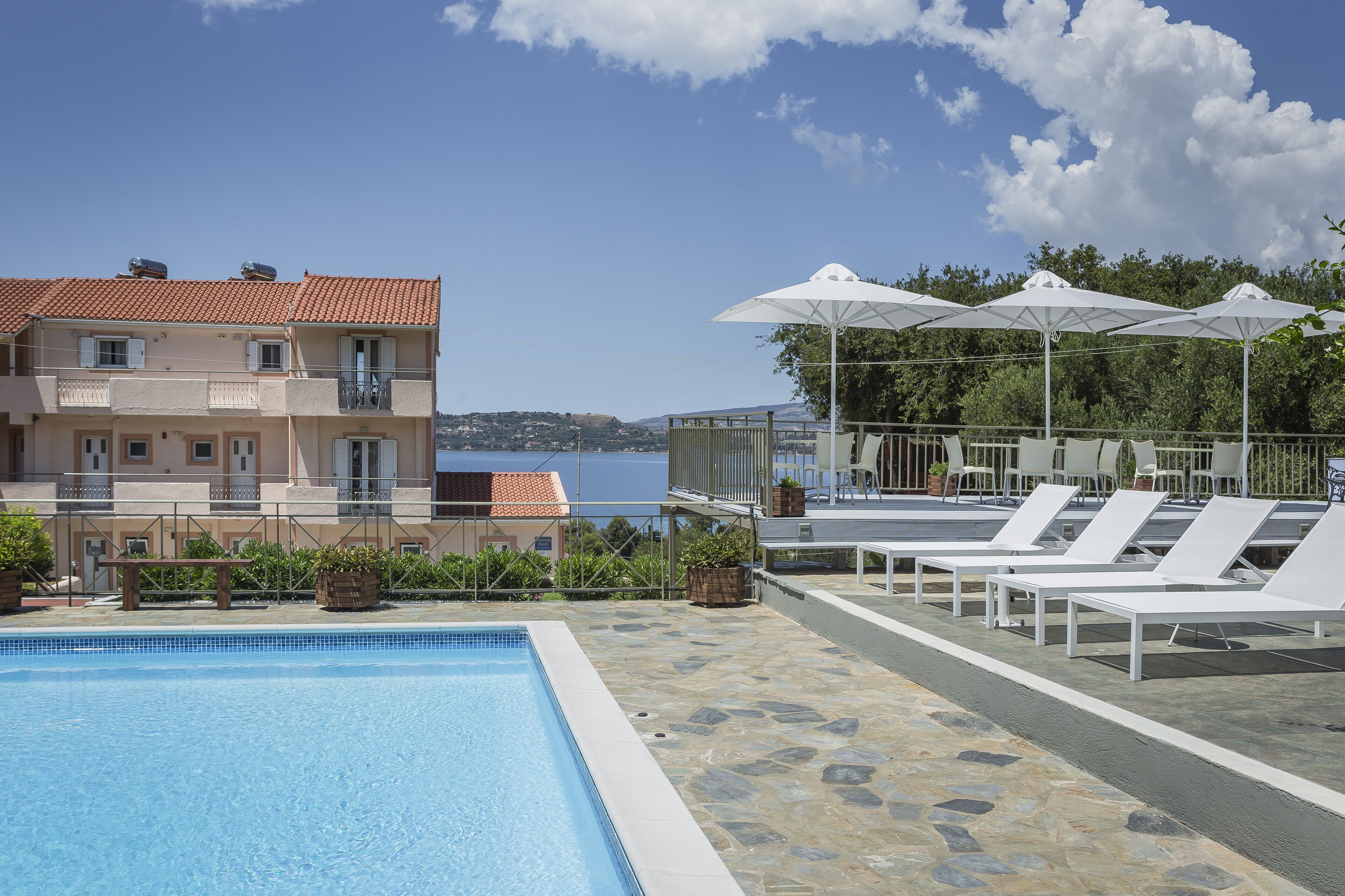 A swimming pool with lounge chairs and umbrellas, a view of a building with a red-tiled roof, and a clear sky.