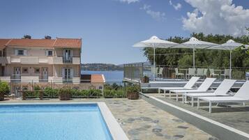 A swimming pool with lounge chairs and umbrellas, a view of a building with a red-tiled roof, and a clear sky.