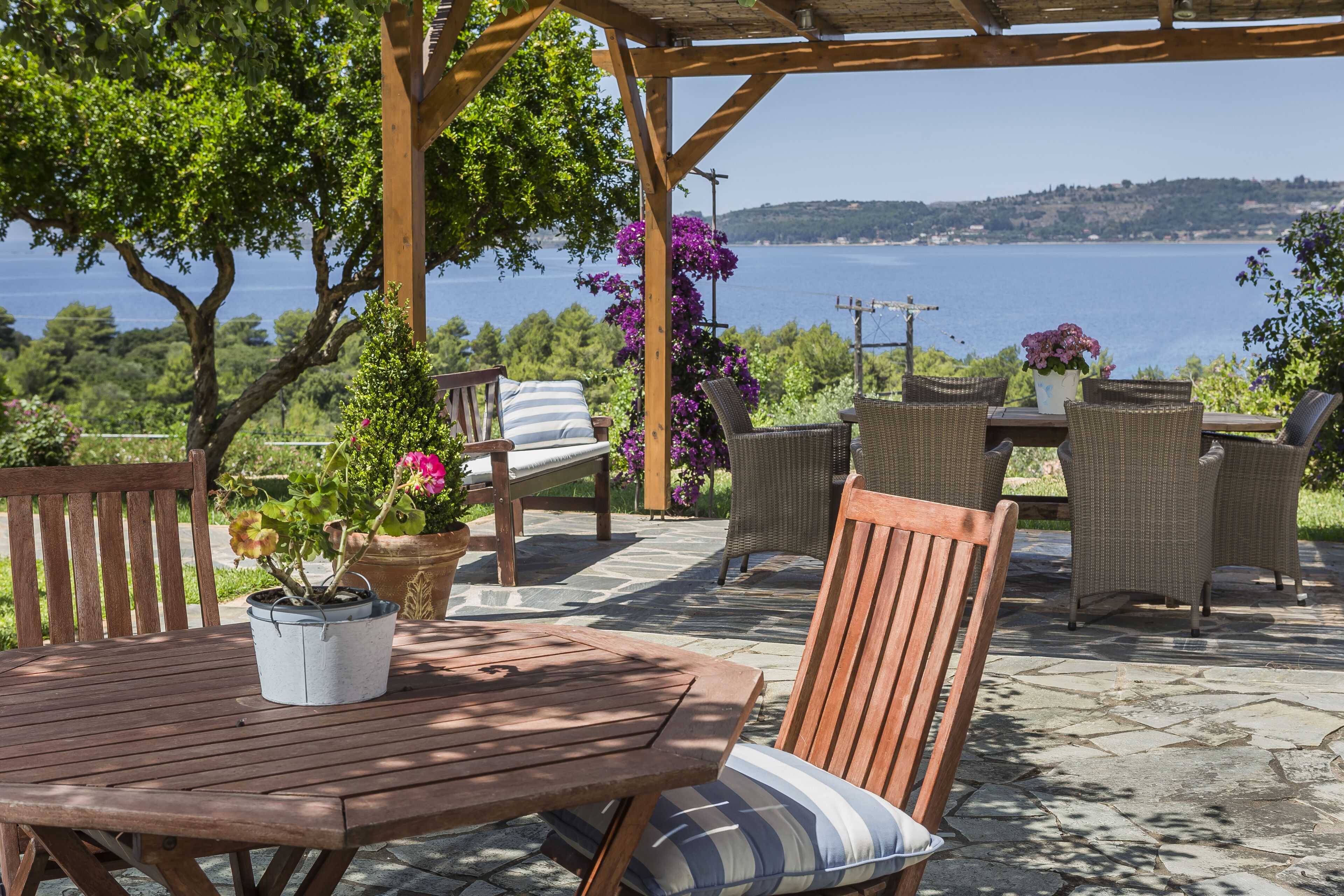 An outdoor patio with a wooden table and chairs, surrounded by potted plants and a view of the sea.