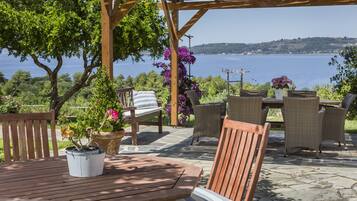 An outdoor patio with a wooden table and chairs, surrounded by potted plants and a view of the sea.