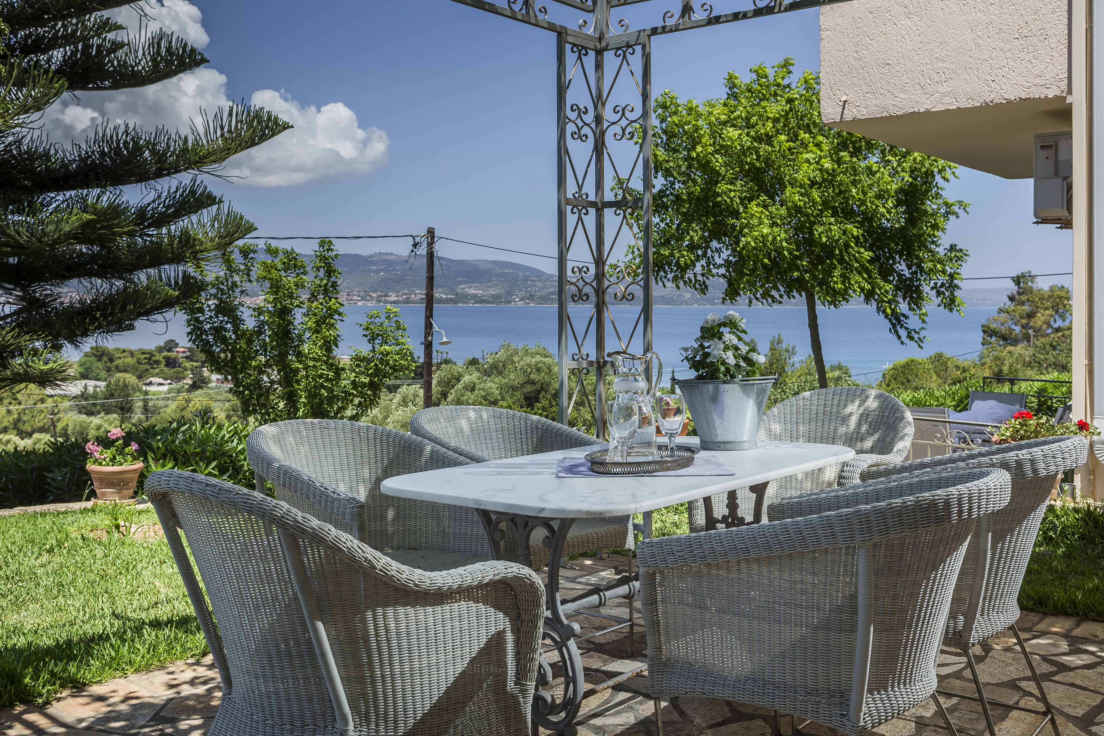 A patio with wicker furniture and a table, overlooking a scenic view of the sea and hills.