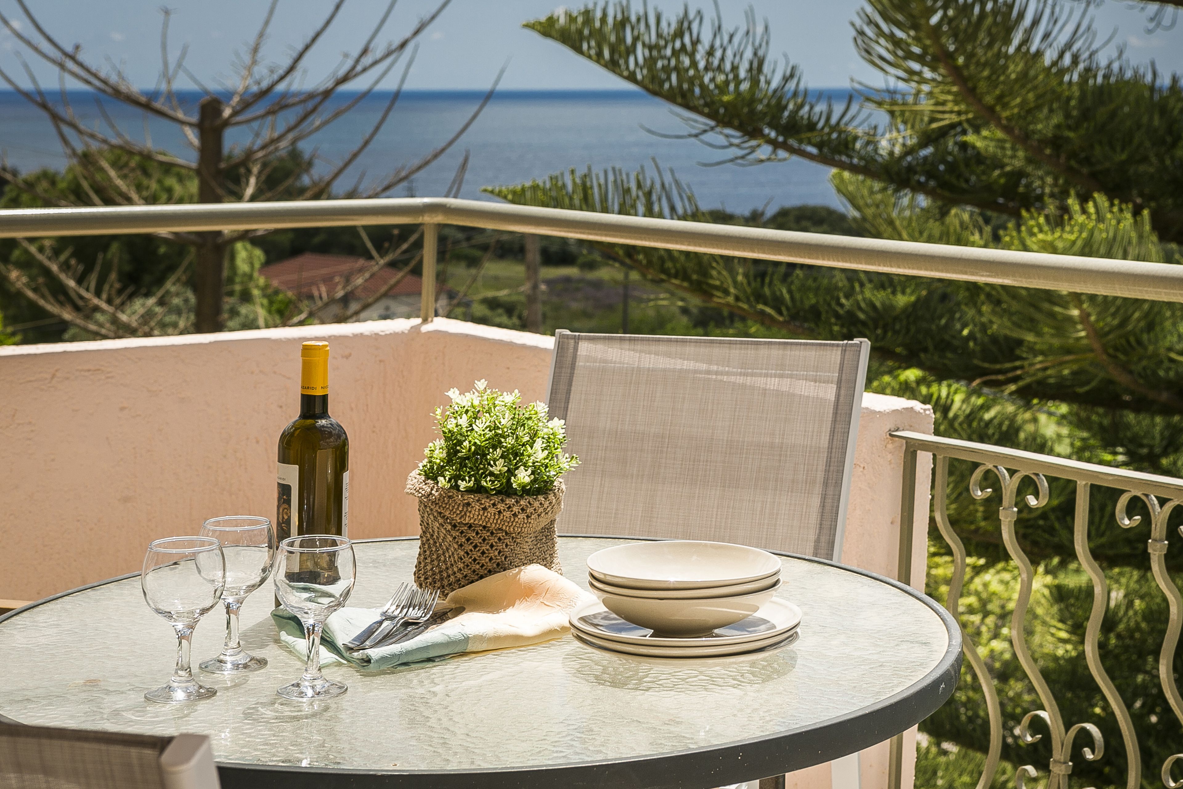 A round glass table with a bottle, glasses, plates, and a small plant on a balcony overlooking the sea.