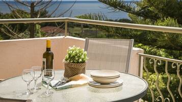 A round glass table with a bottle, glasses, plates, and a small plant on a balcony overlooking the sea.