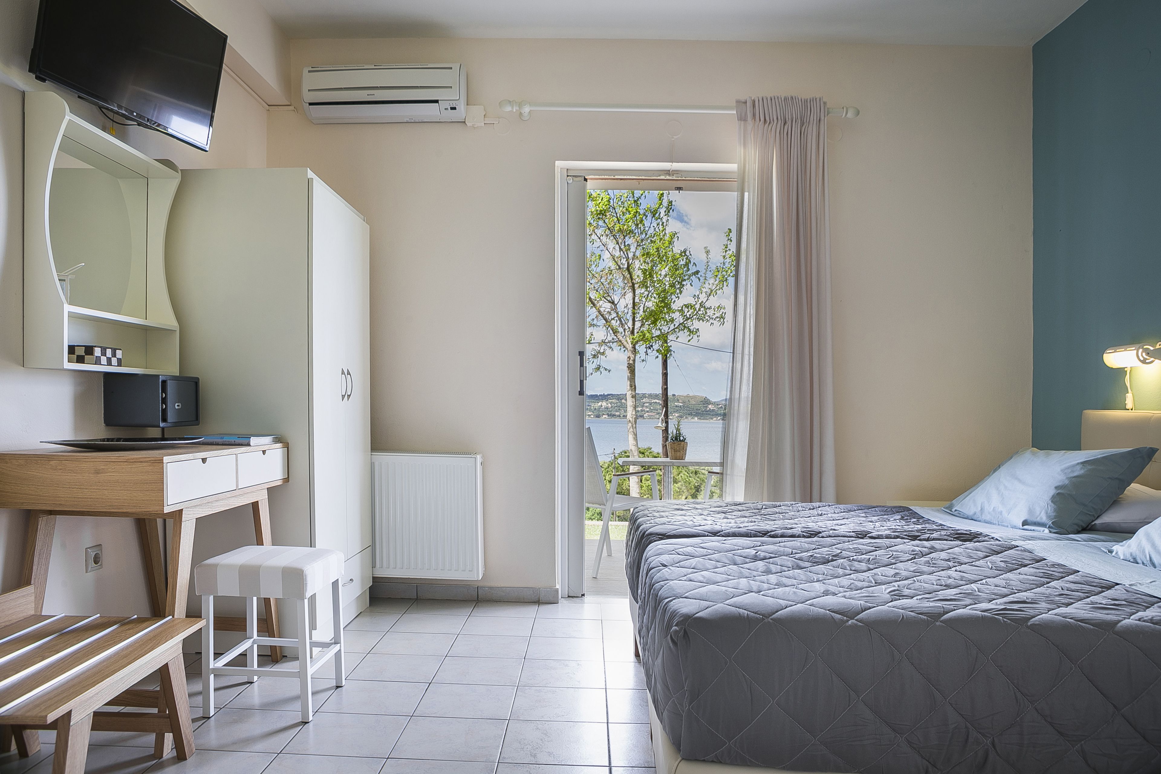 Un dormitorio con cama, escritorio con computadora, silla y ventana con vista a árboles y una masa de agua.