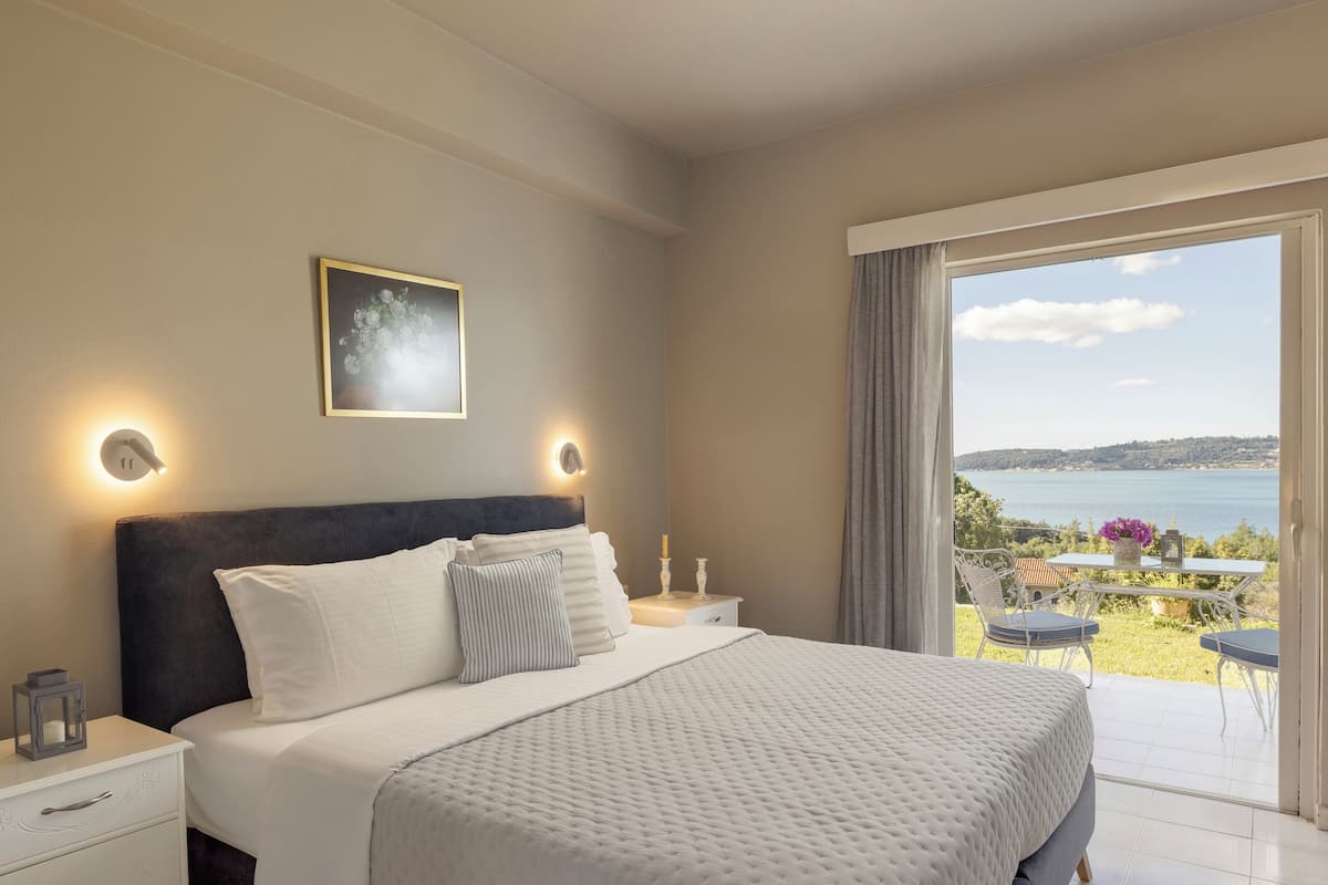 Een slaapkamer met een bed, nachtkastjes, een raam met uitzicht op zee en een balkon met buitenmeubilair.