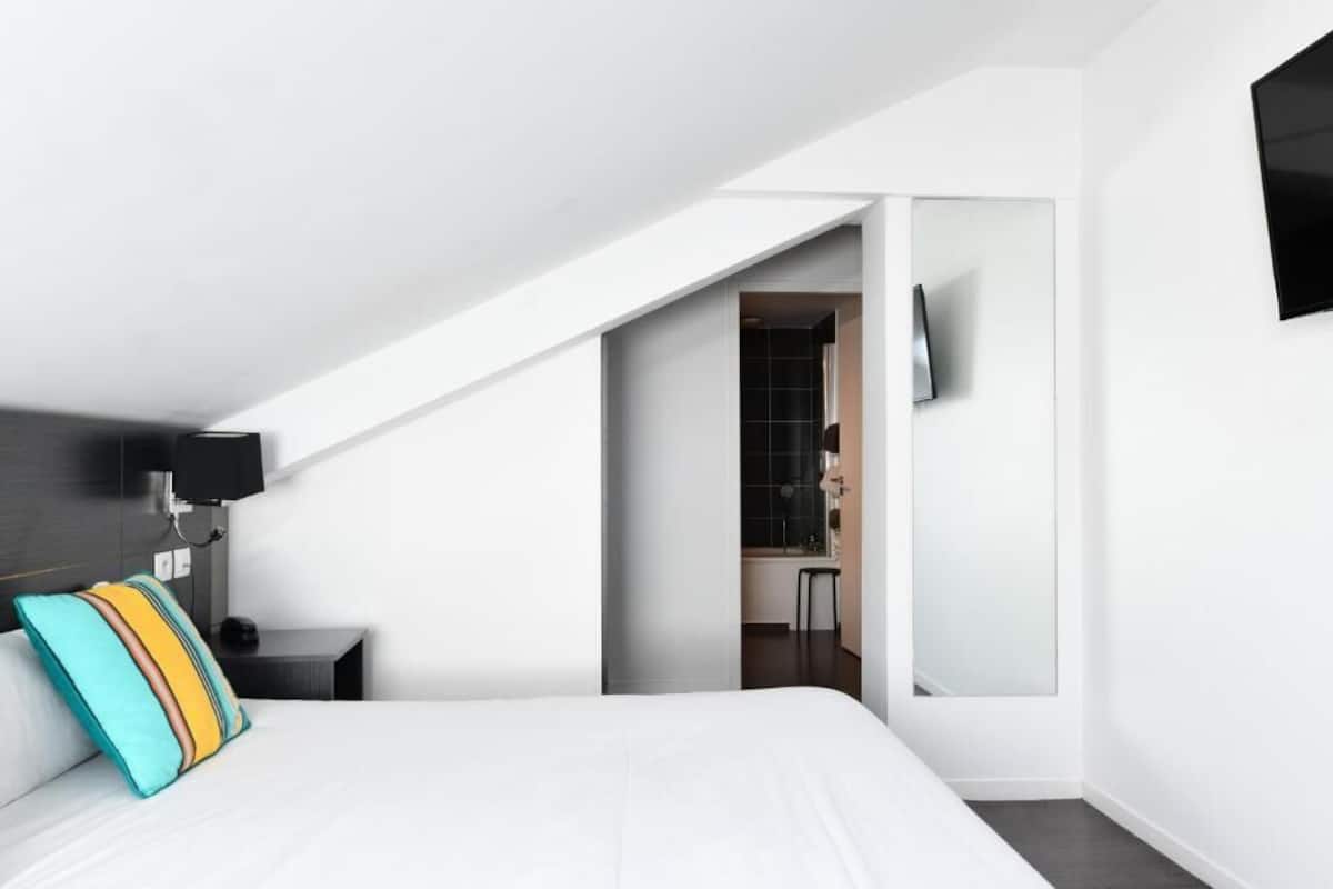 A modern hotel room with a bed, a nightstand, a TV mounted on the wall, and a bathroom visible through an open door.