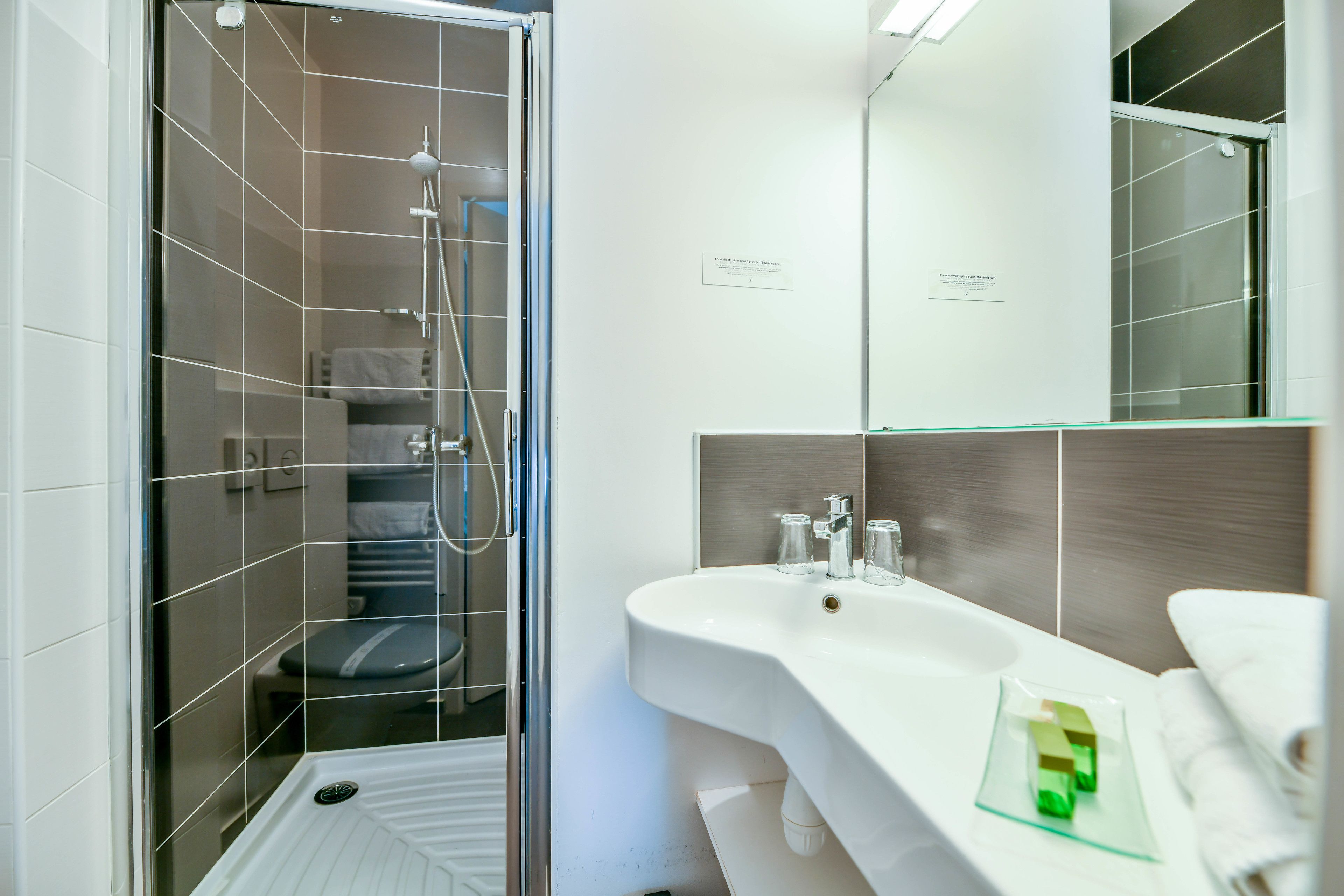 Un baño moderno con ducha de vidrio, lavamanos blanco con jabonera de vidrio y un espejo grande sobre el lavamanos.