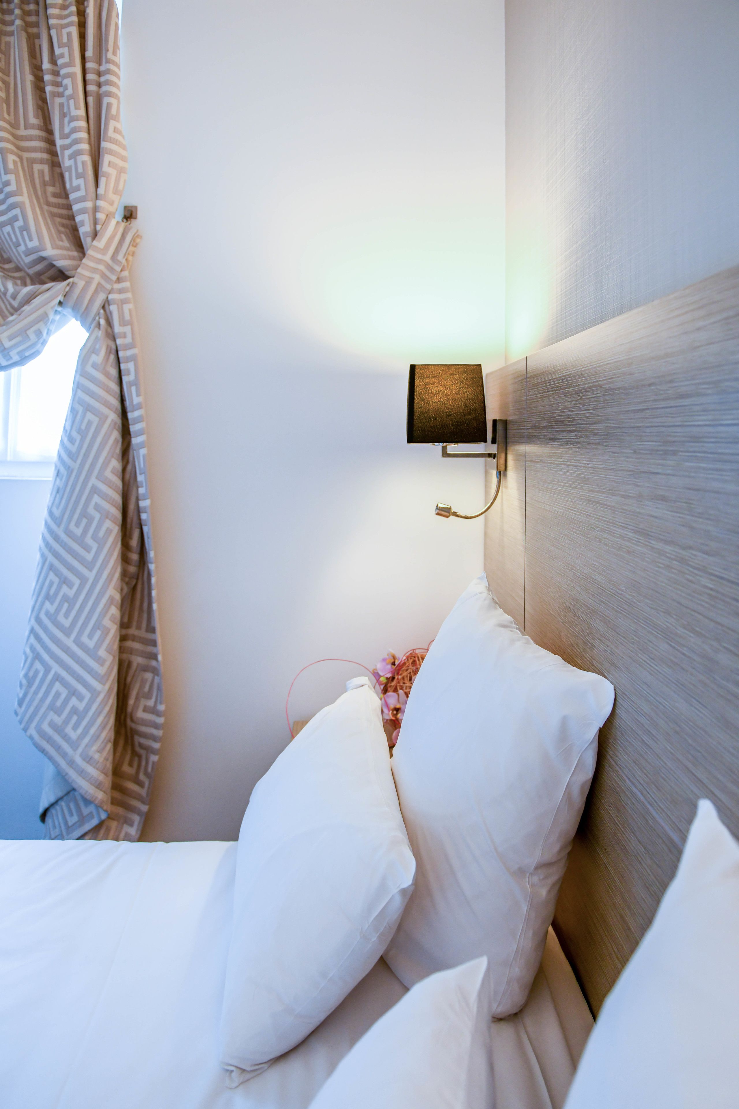 A neatly made bed with white linens and pillows, a wall-mounted light fixture, and a window with curtains.