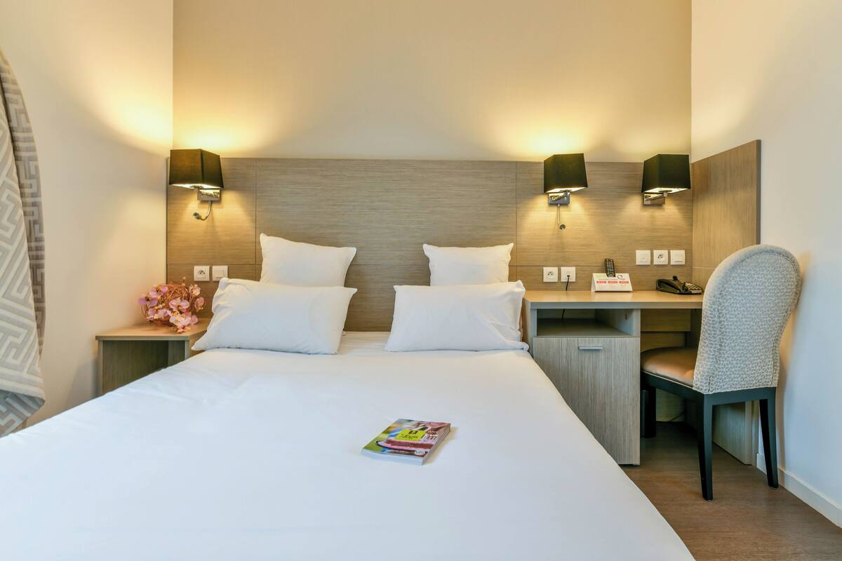 A hotel room with a large bed, two bedside tables with lamps, a desk with a chair, and a small nightstand with a vase of flowers.