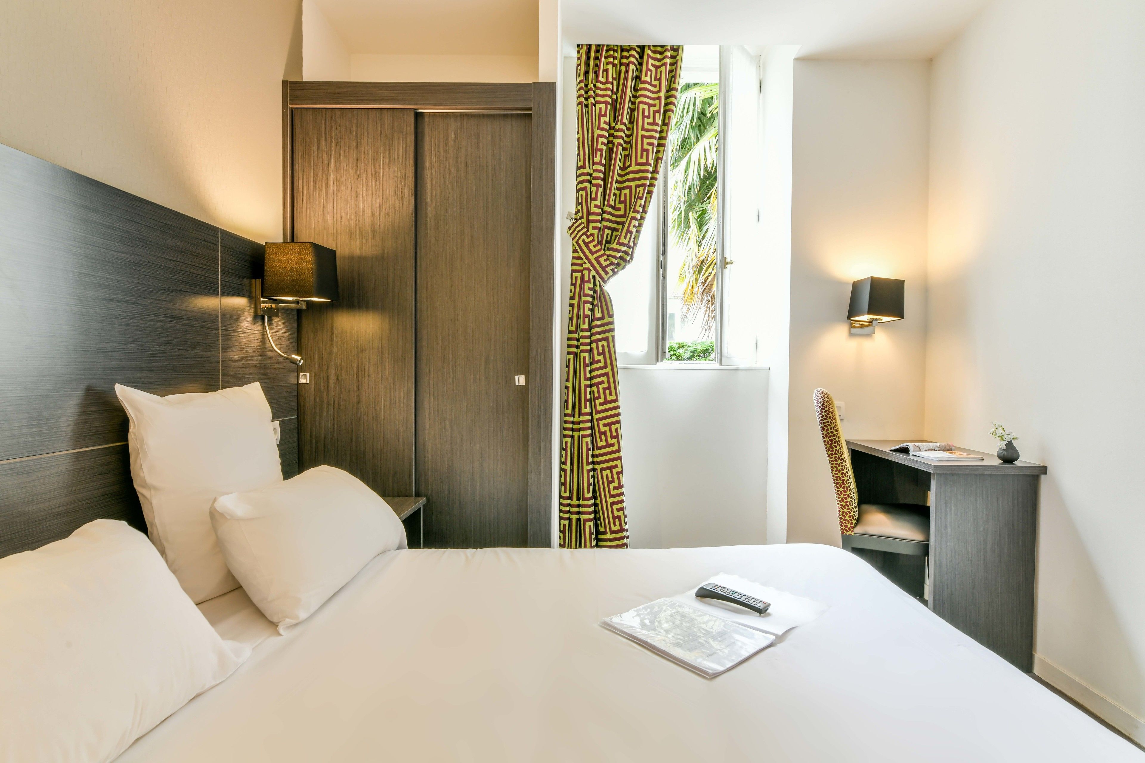 A hotel room with a bed, a desk with a chair, a wardrobe, and two wall-mounted lamps.