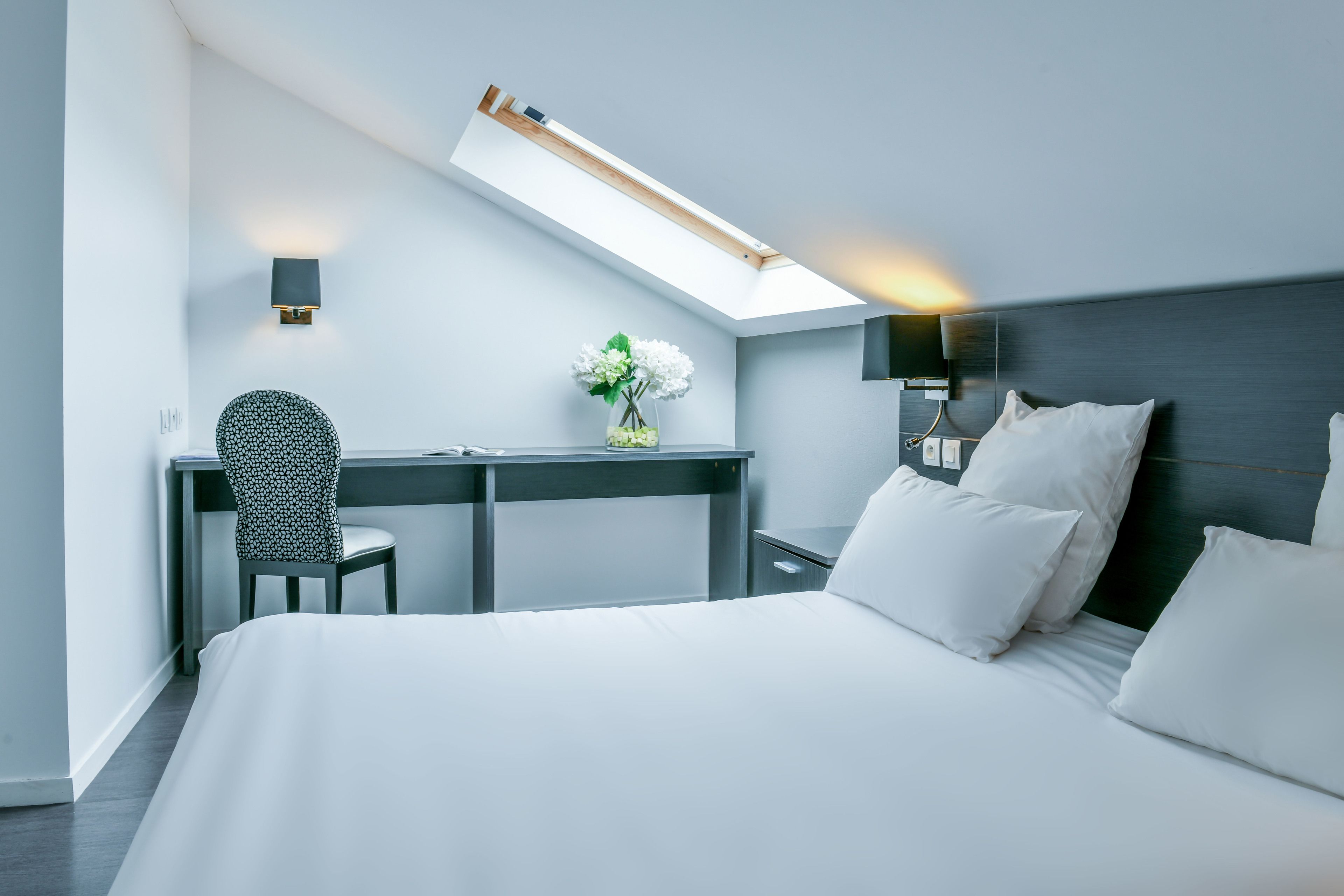 Een moderne hotelkamer met dakraam, bureau, stoel, bed met witte beddengoed en nachtkastje met lamp.