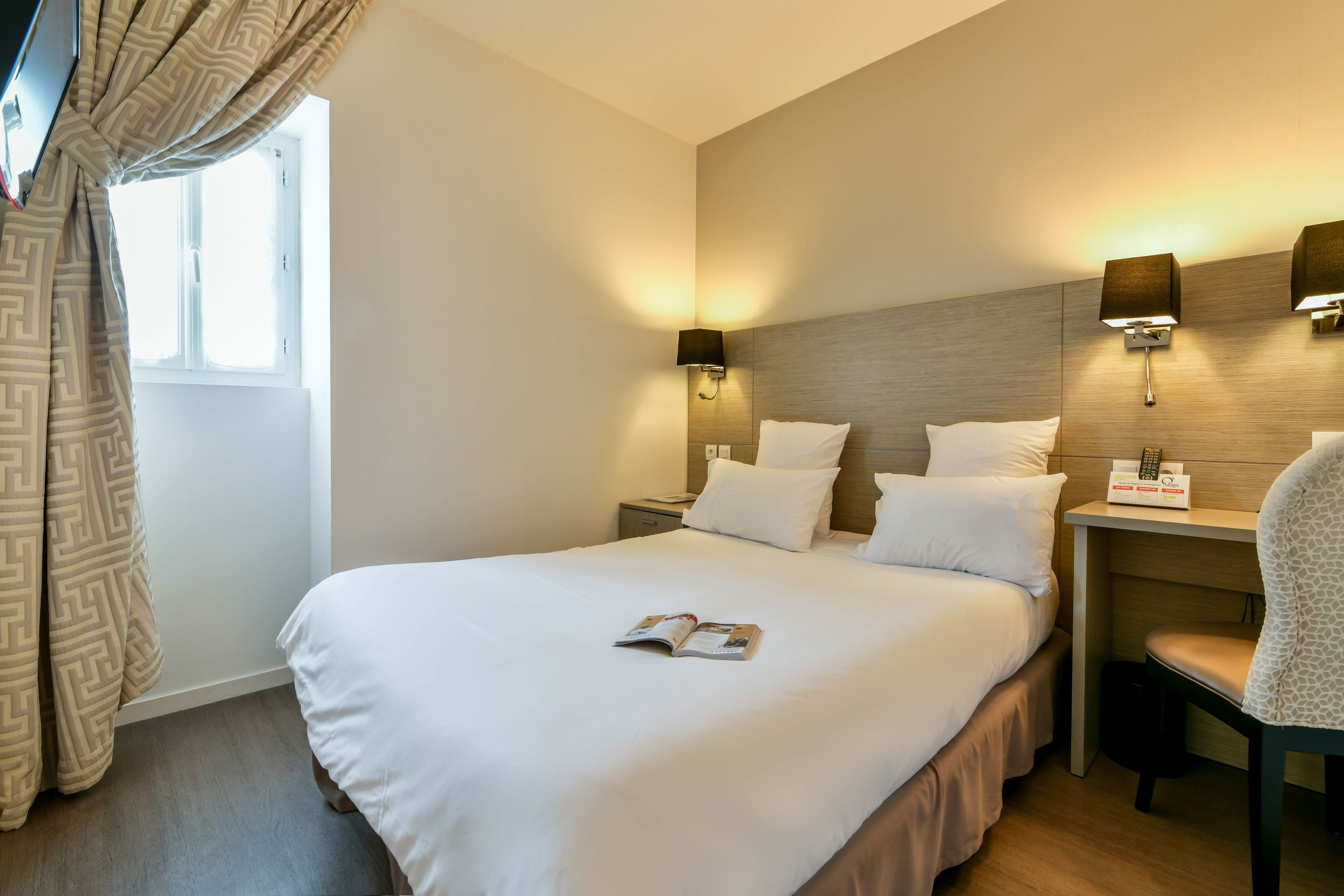 A neatly made bed with a magazine, two bedside tables with lamps, a chair, and a window with curtains.