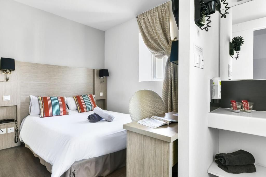 A hotel room with a bed, a desk, a chair, a bedside table, a lamp, a mirror, a shelf with toiletries, and a window with curtains.