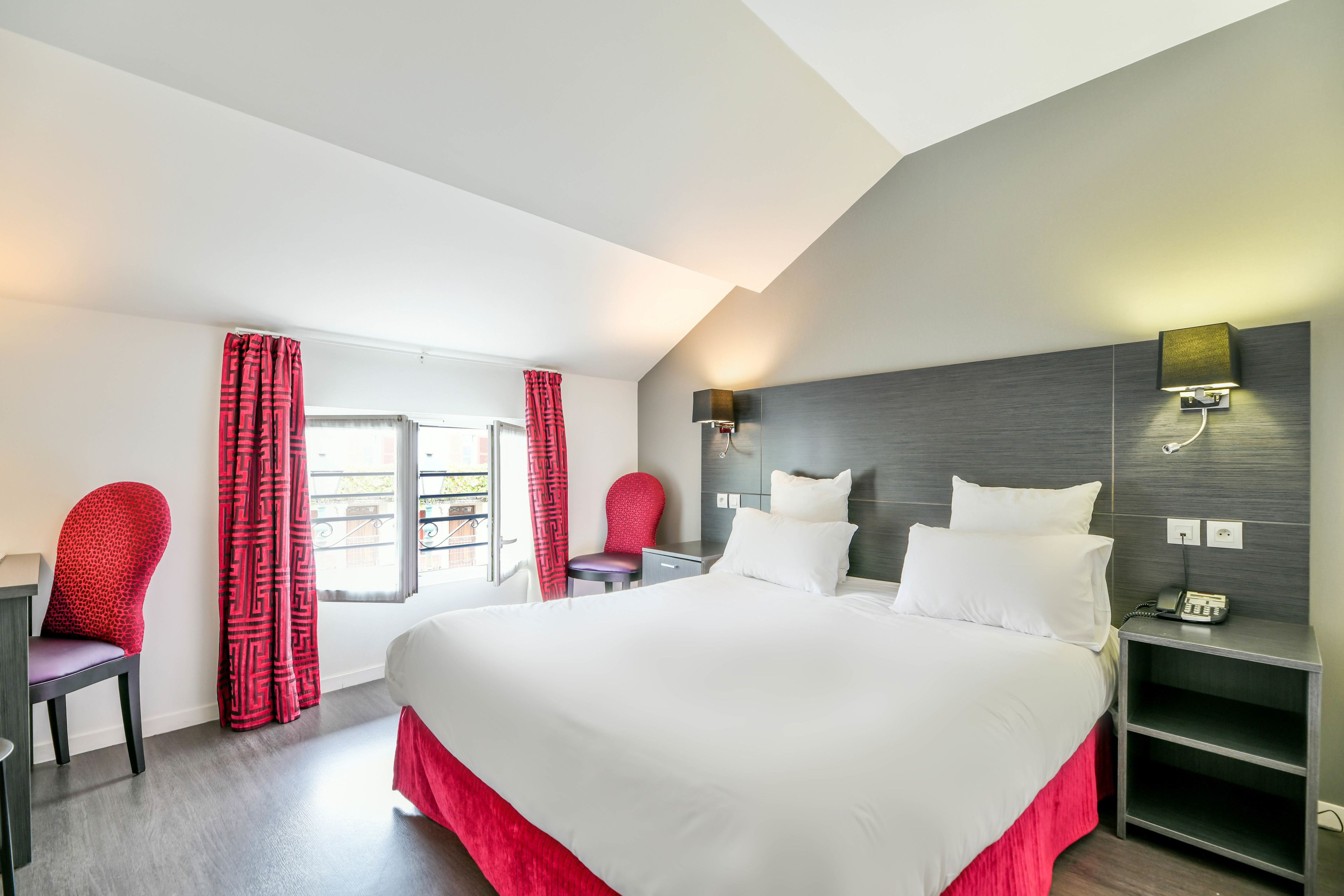 A modern hotel room with a large bed, a desk, and a chair. There is a window with curtains, a nightstand with a lamp, and a phone.