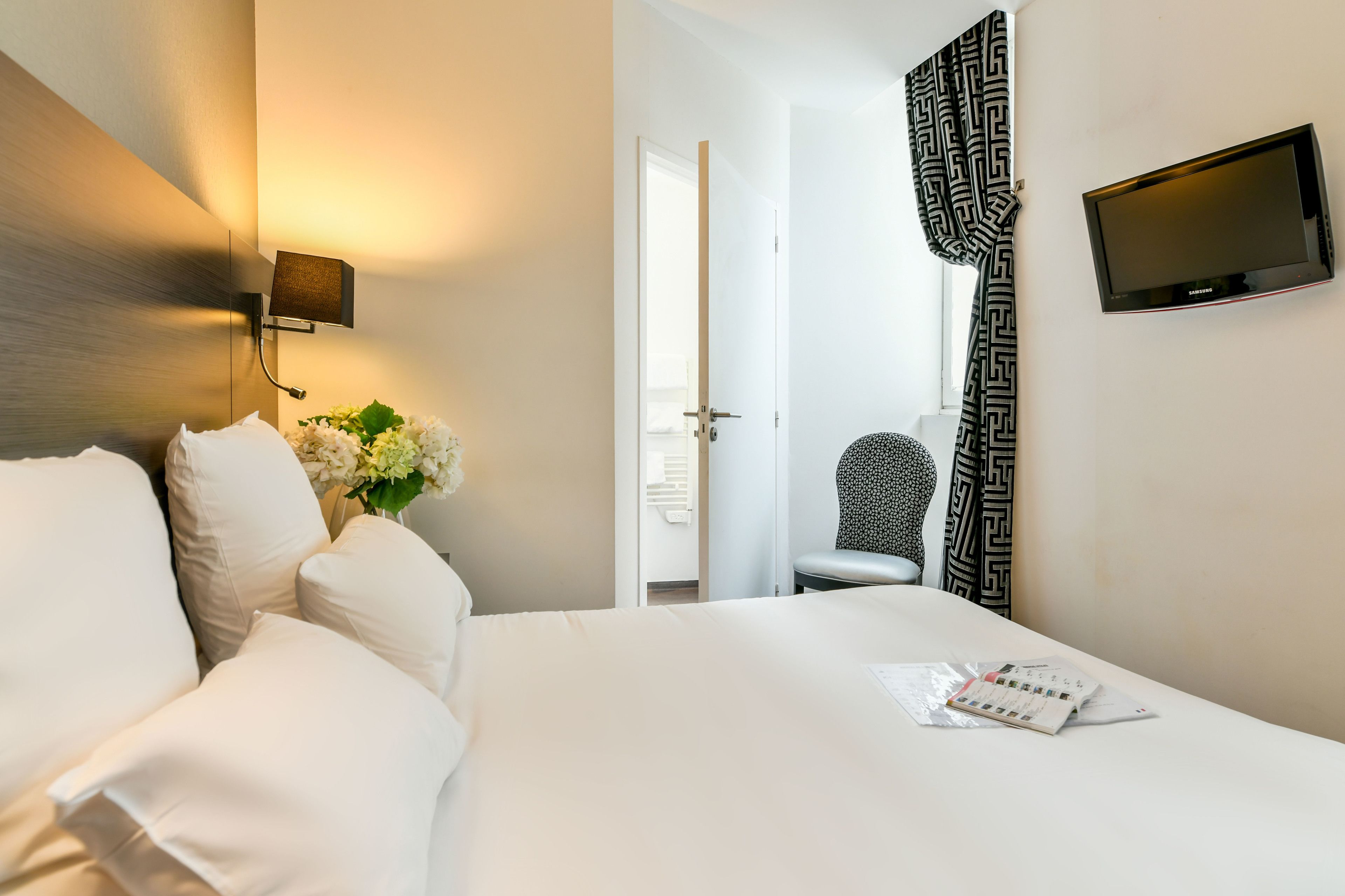A hotel room with a bed, a wall-mounted TV, a chair, and a door leading to another room.