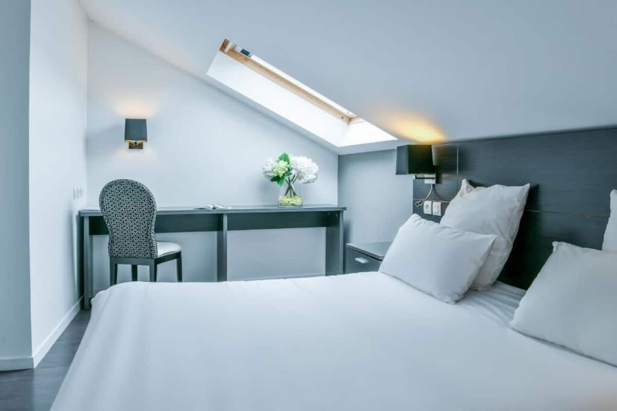 A modern hotel room with a bed, desk, and chair, a skylight, and a wall-mounted lamp.