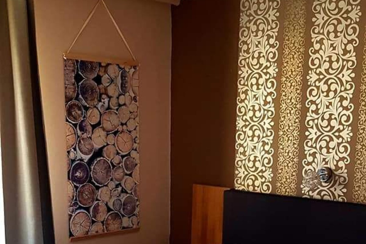 Un lit bien fait, avec du linge de lit blanc et un plaid beige, une œuvre d’art représentant des troncs d’arbres accrochée au mur, et un rideau à motifs.