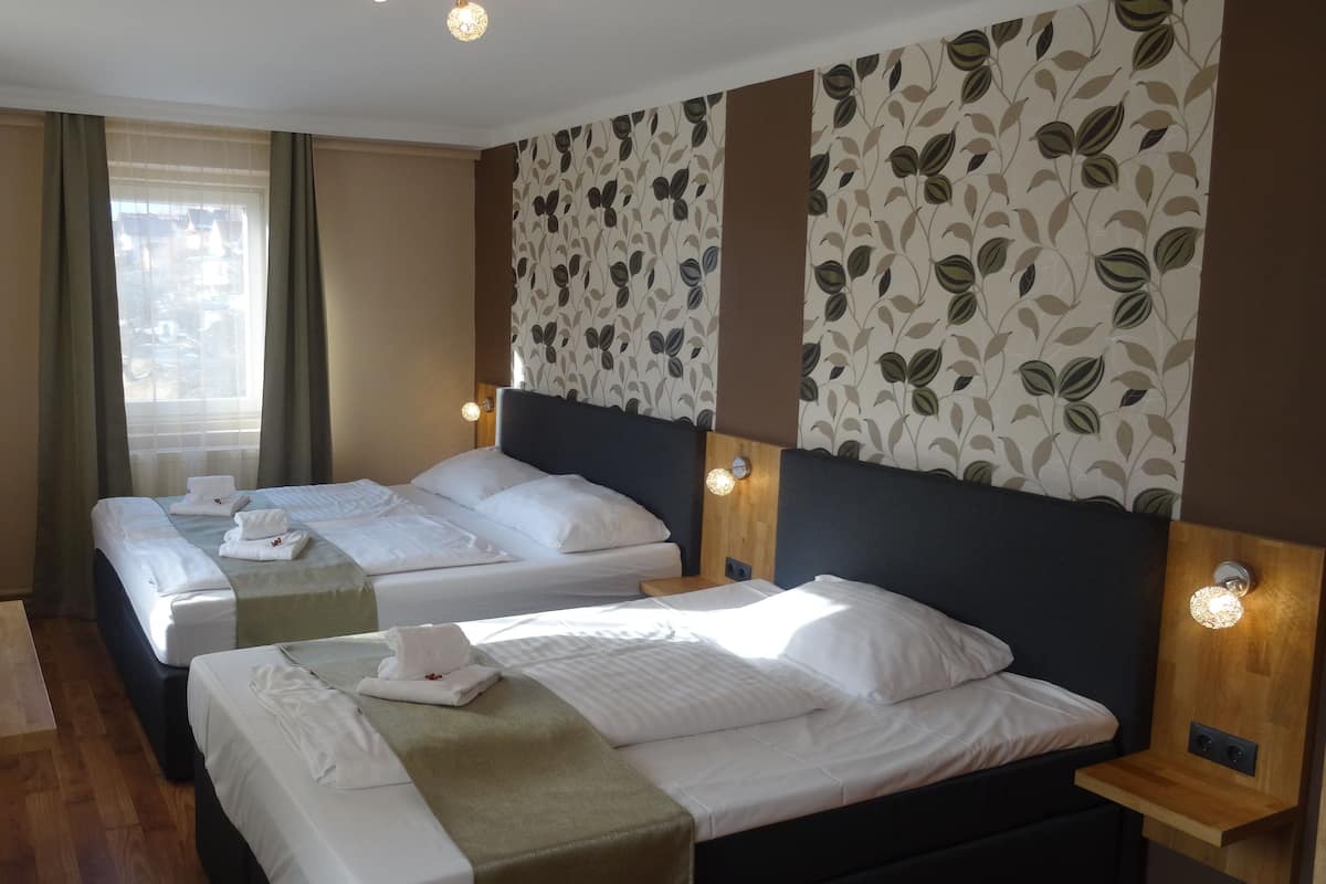Ein Hotelzimmer mit zwei Betten, einem Holztisch und Blick durch ein mit Vorhängen versehenes Fenster nach draußen.