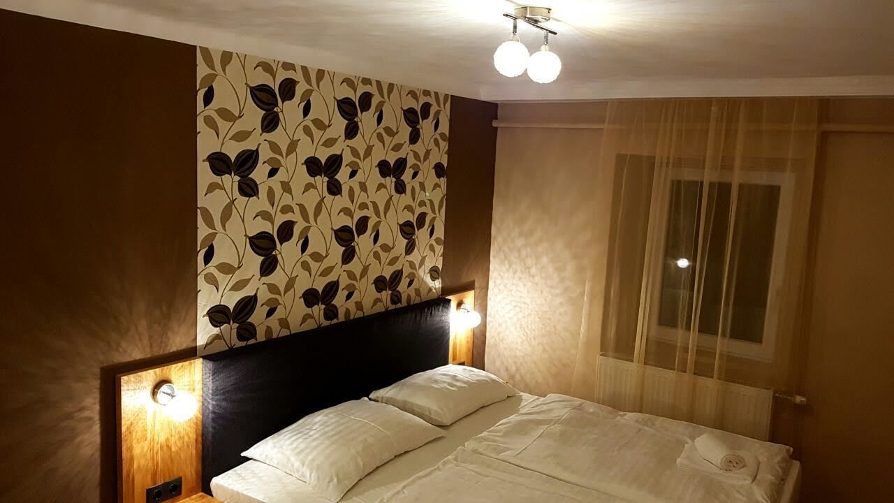 Une chambre d’hôtel avec un lit, une tête de lit ornée d’un motif de feuilles, deux lampes de chevet et une fenêtre avec des rideaux légers.