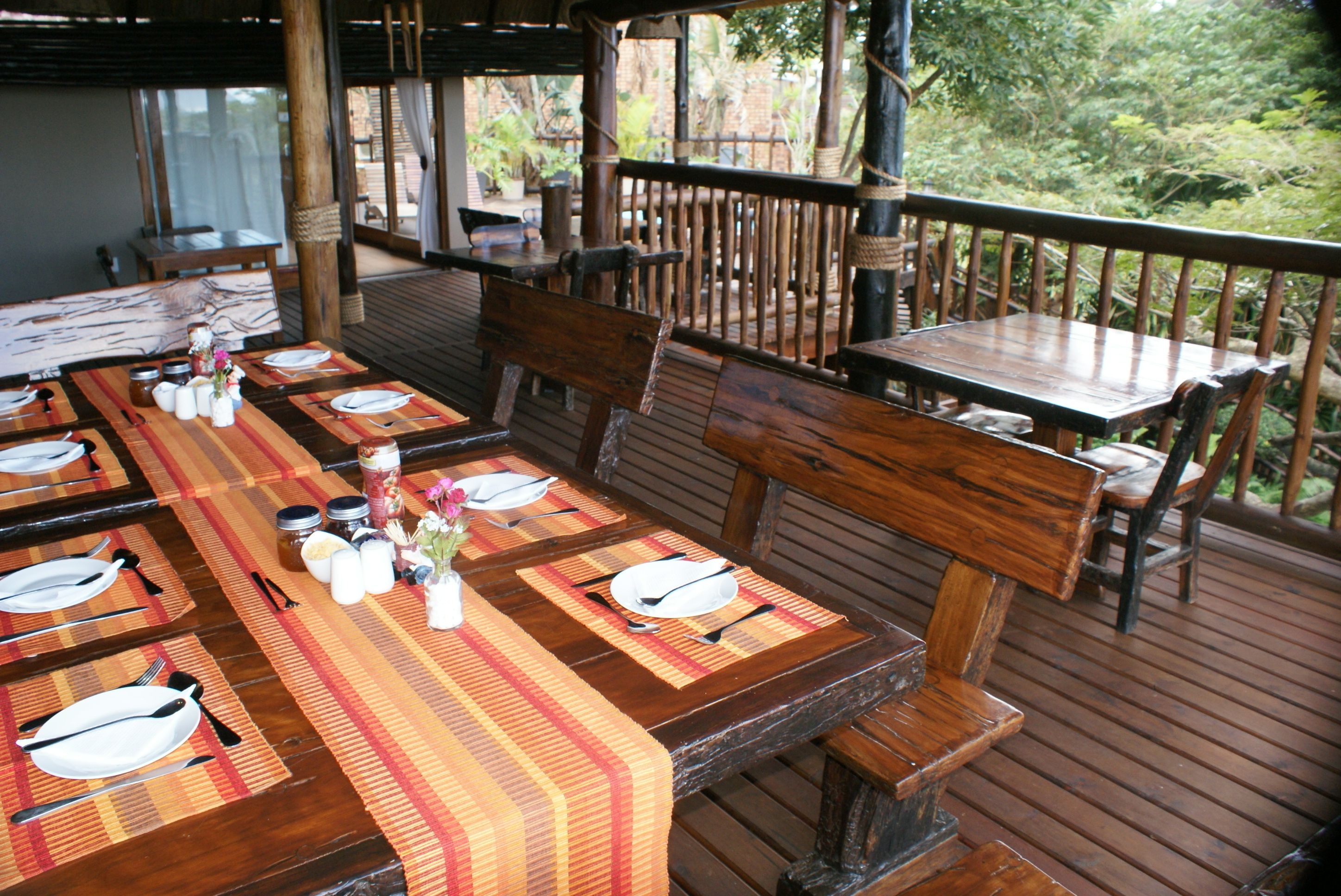 A wooden deck with a long dining table set for a meal, surrounded by wooden chairs and benches.