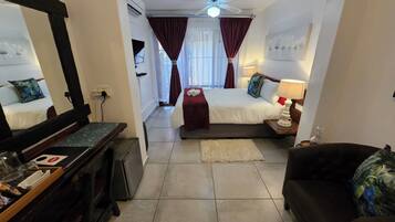 A hotel room with a bed, a desk, a chair, a mirror, a window with curtains, a lamp, and a painting on the wall.
