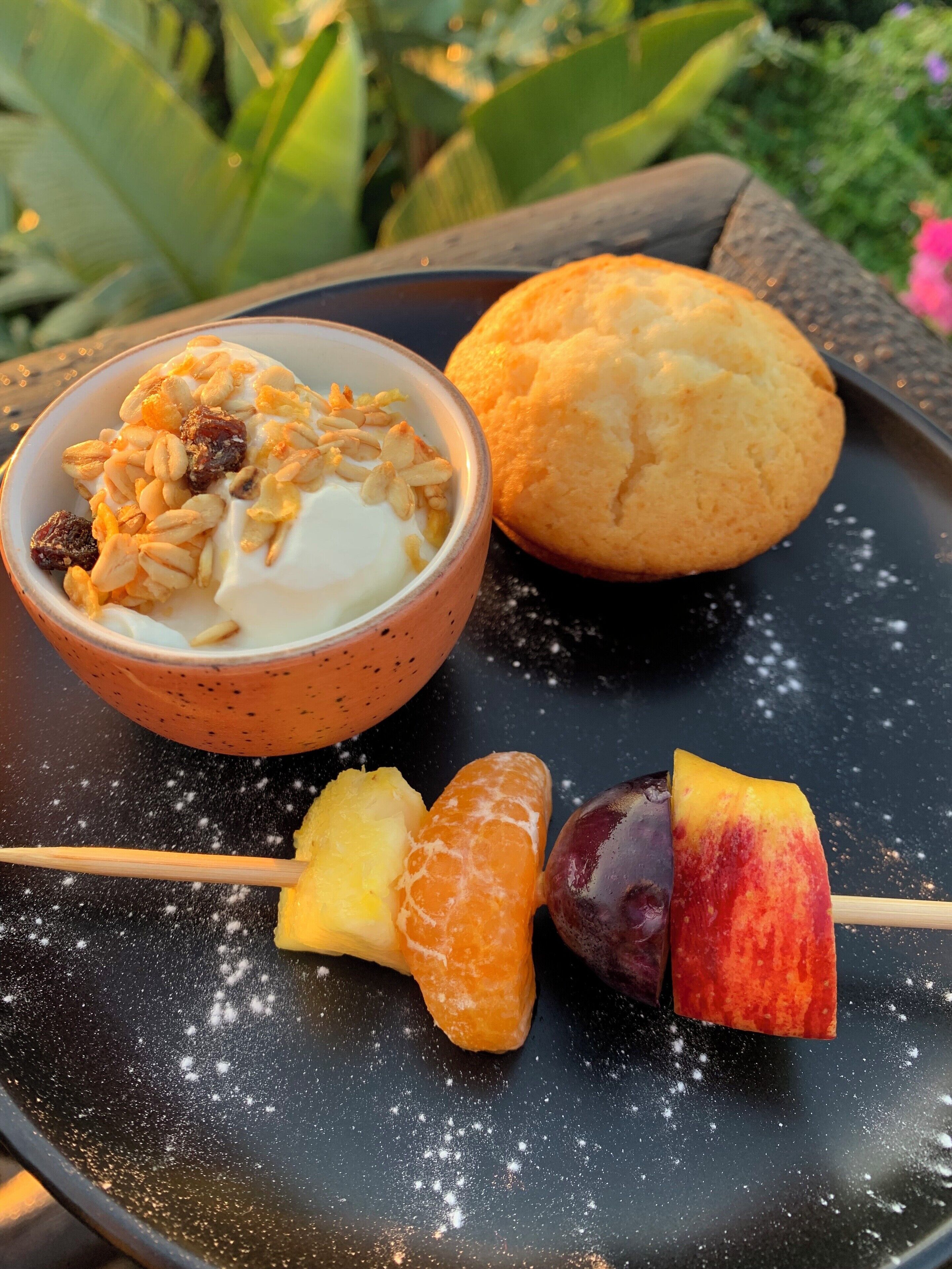 A plate with a bowl of yogurt topped with granola and almonds, a muffin, and fruit skewers.