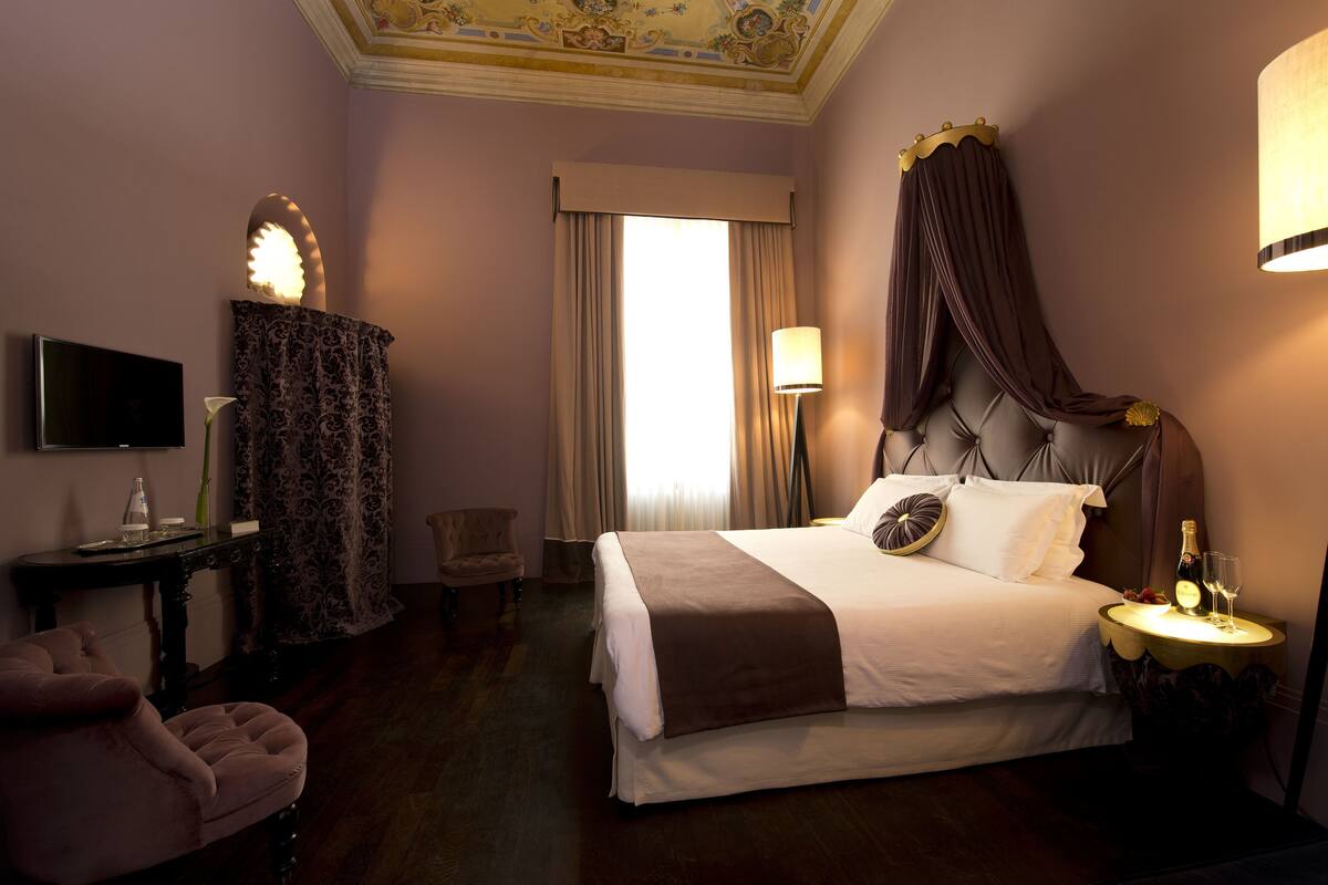 A hotel room with a large bed, a television, a side table with a bottle and glasses, a chair, and a window with curtains.