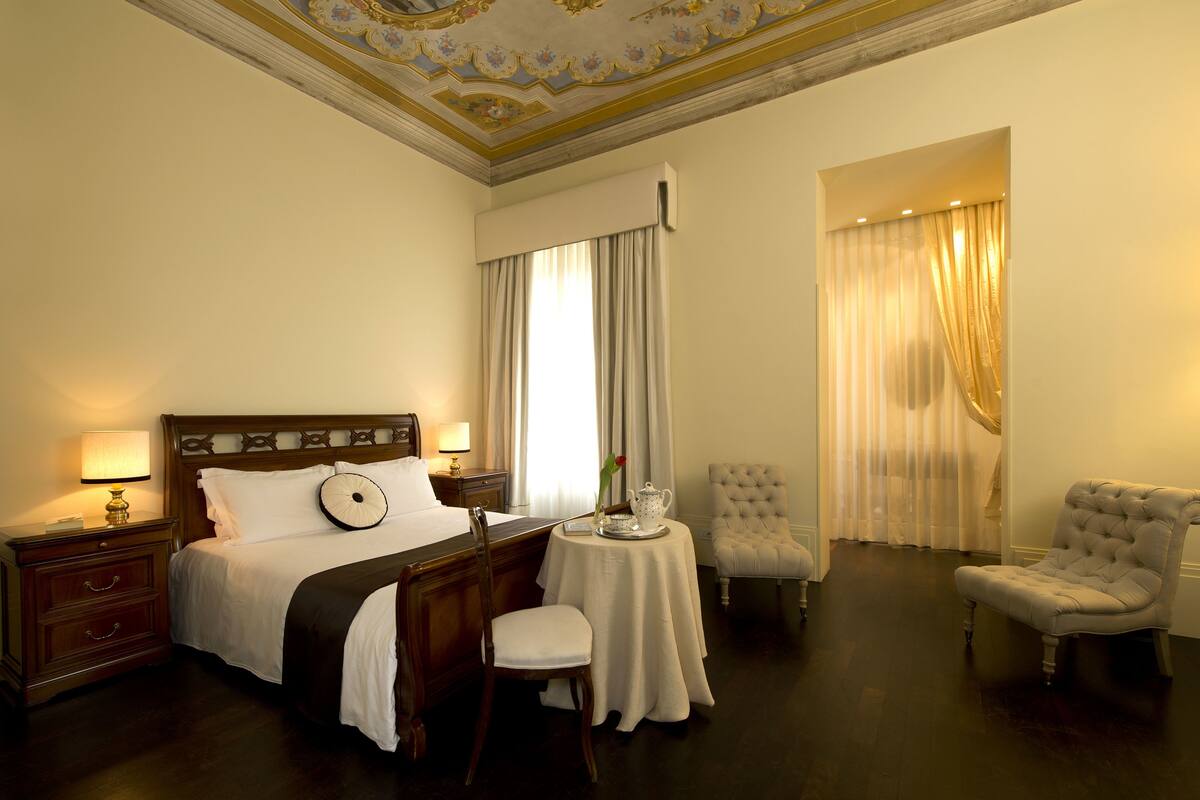 A hotel room with a large bed, a bedside table with a lamp, a small round table with a teapot, two chairs, and a large window with curtains.
