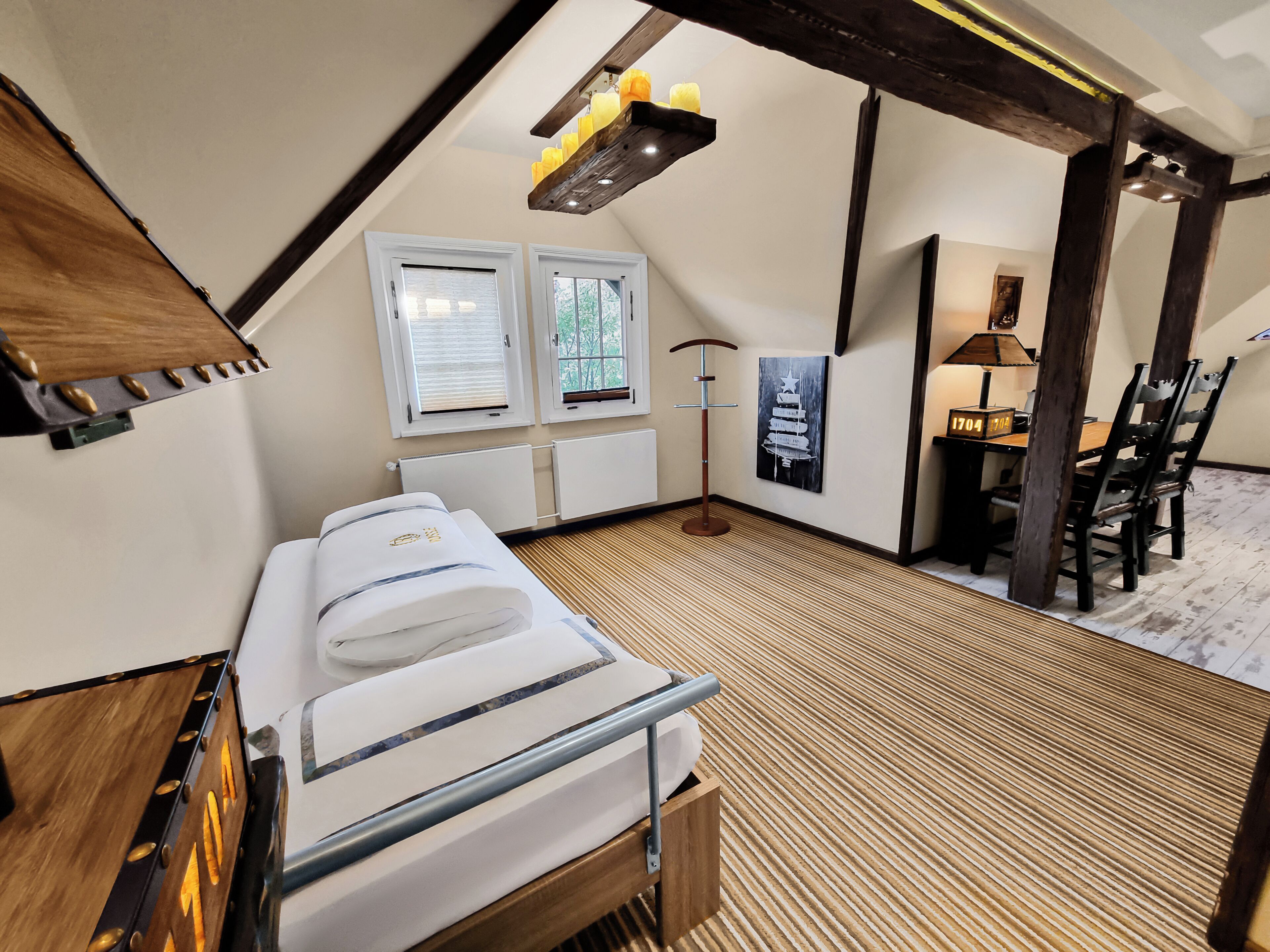 Une pièce confortable avec un lit, un bureau et une chaise. Il y a des fenêtres avec des stores, une lampe et une étagère fixée au mur où sont posées des bougies.