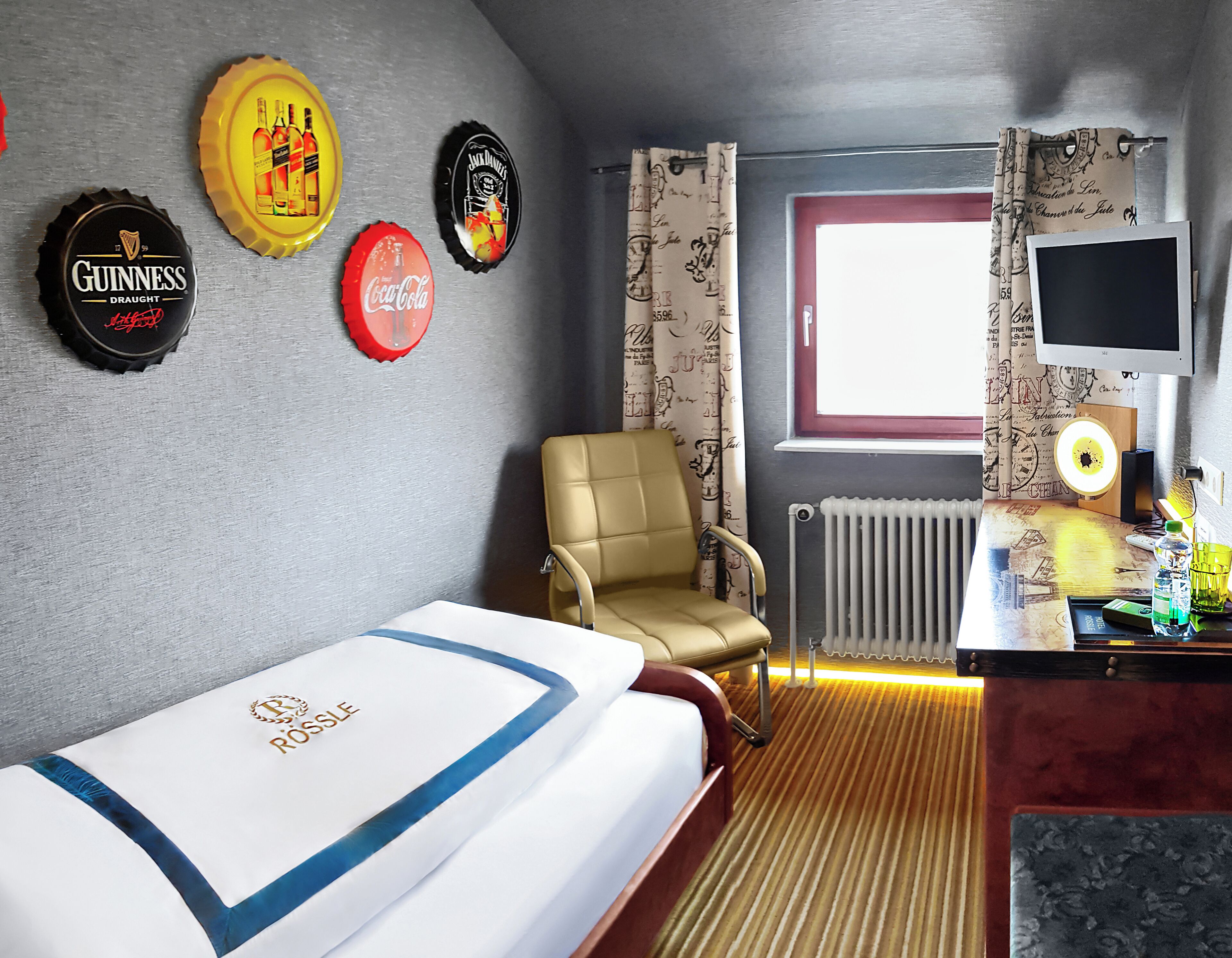 A hotel room with a bed, a chair, a desk with a computer, and wall decorations including bottle caps.